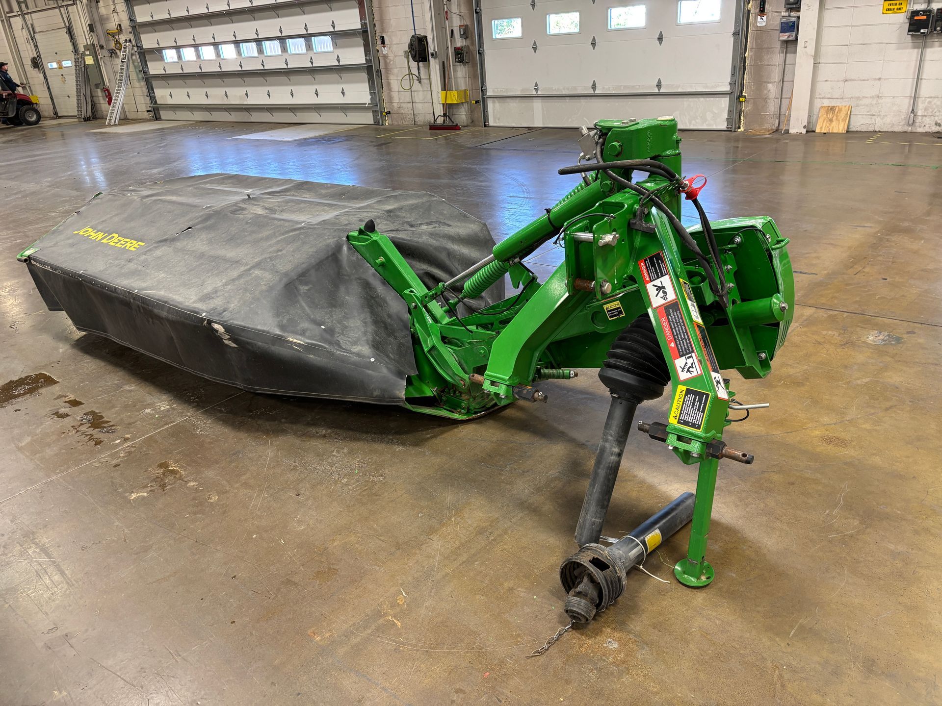 Green, covered John Deere disc mower on a concrete floor in a warehouse setting, front angled shot.