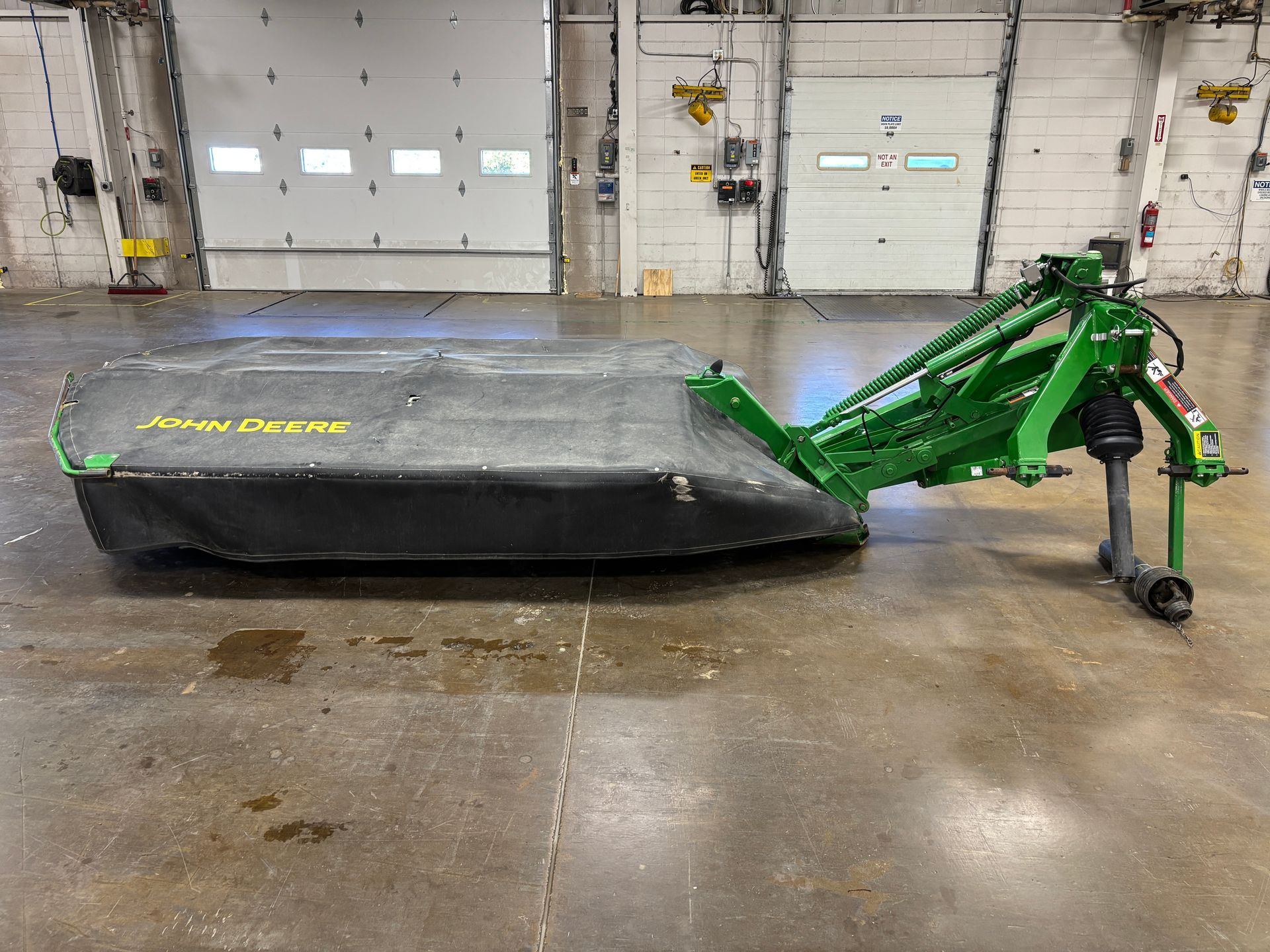Green, covered John Deere disc mower on a concrete floor in a warehouse setting, side shot.