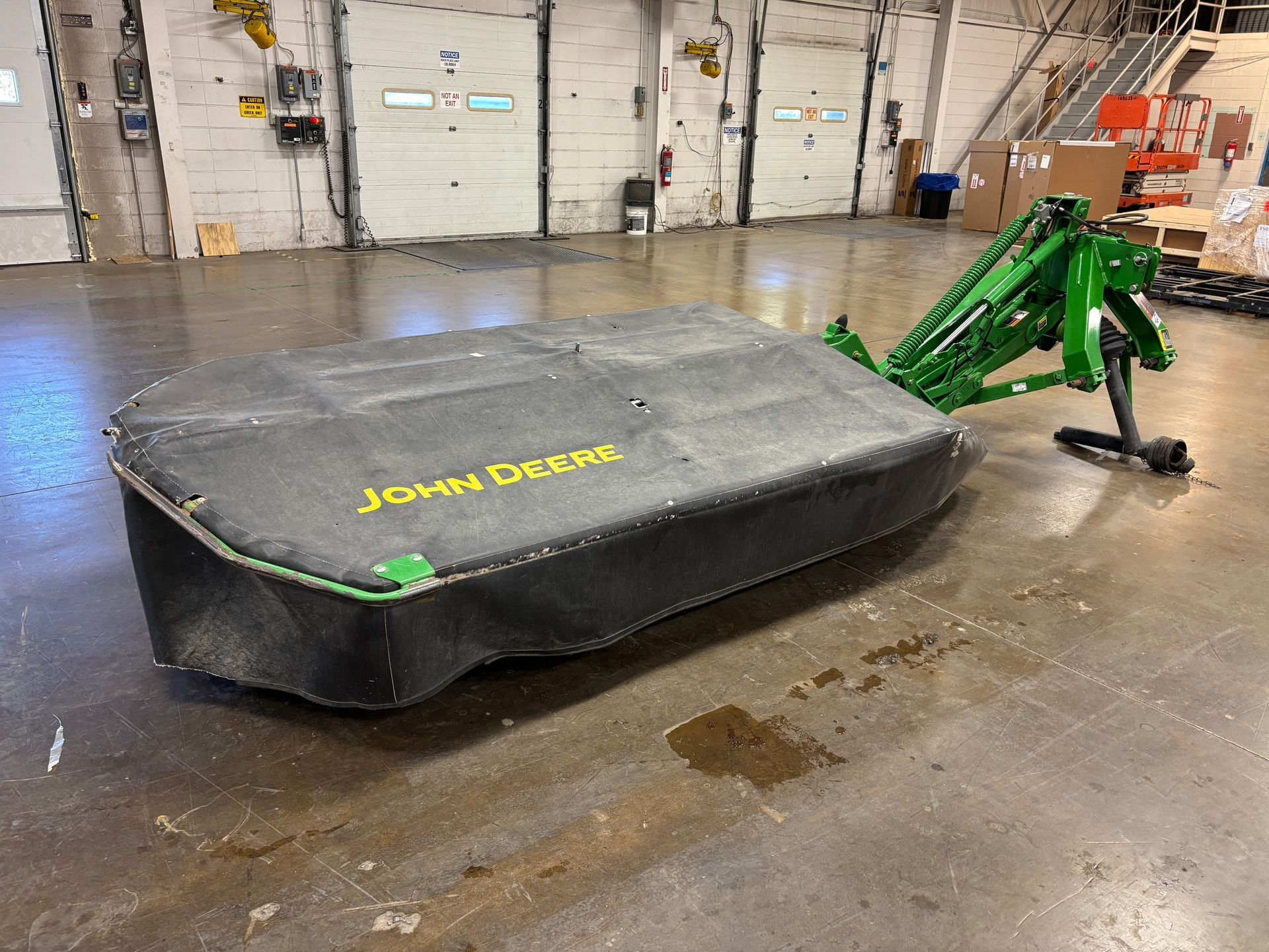 Green, covered John Deere disc mower on a concrete floor in a warehouse setting, angled shot.