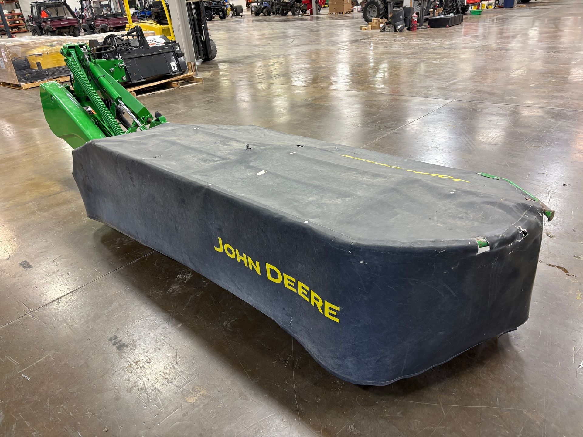 Green, covered John Deere disc mower on a concrete floor in a warehouse setting, back angled shot.