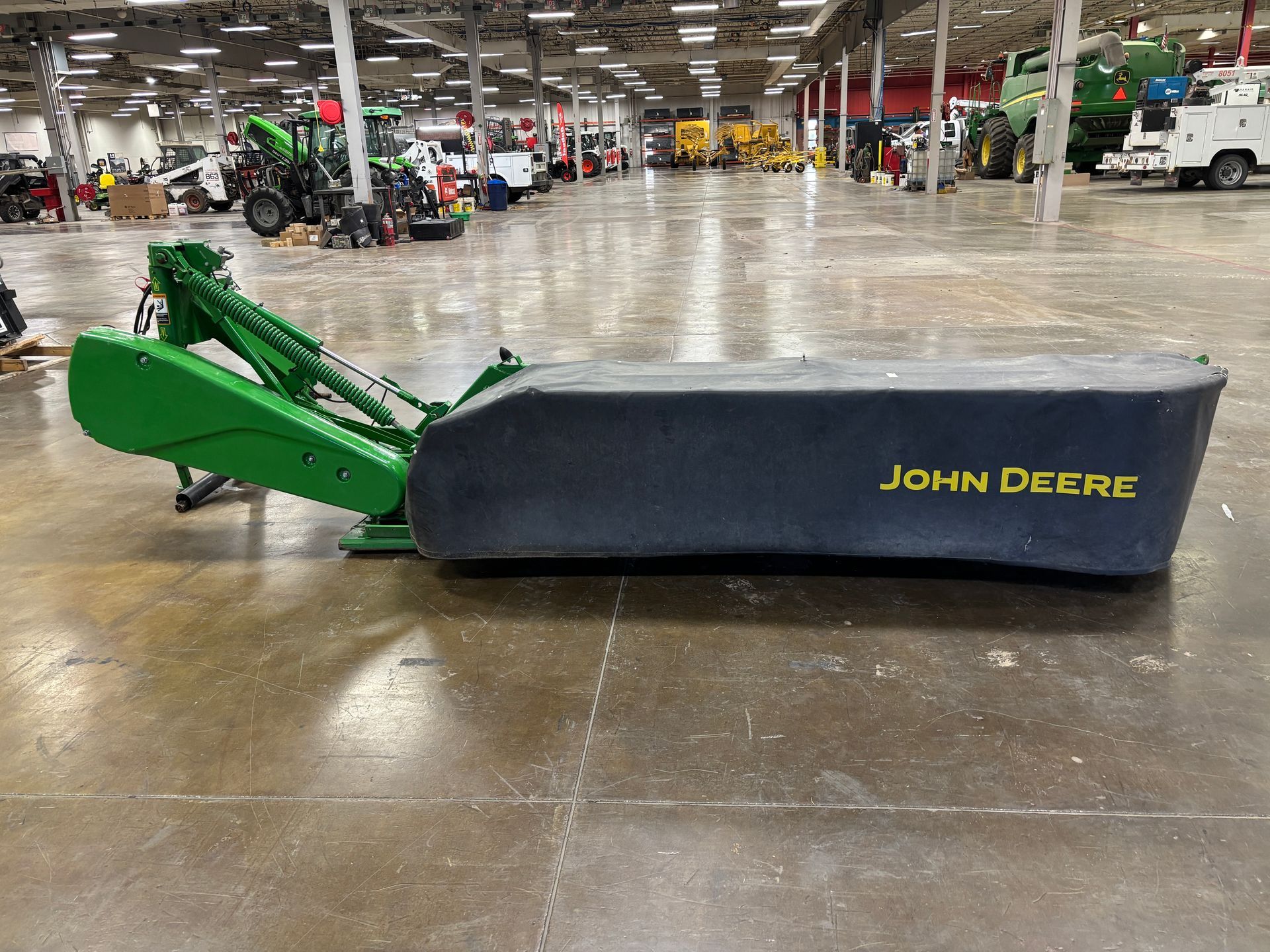 Green, covered John Deere disc mower on a concrete floor in a warehouse setting, side shot.