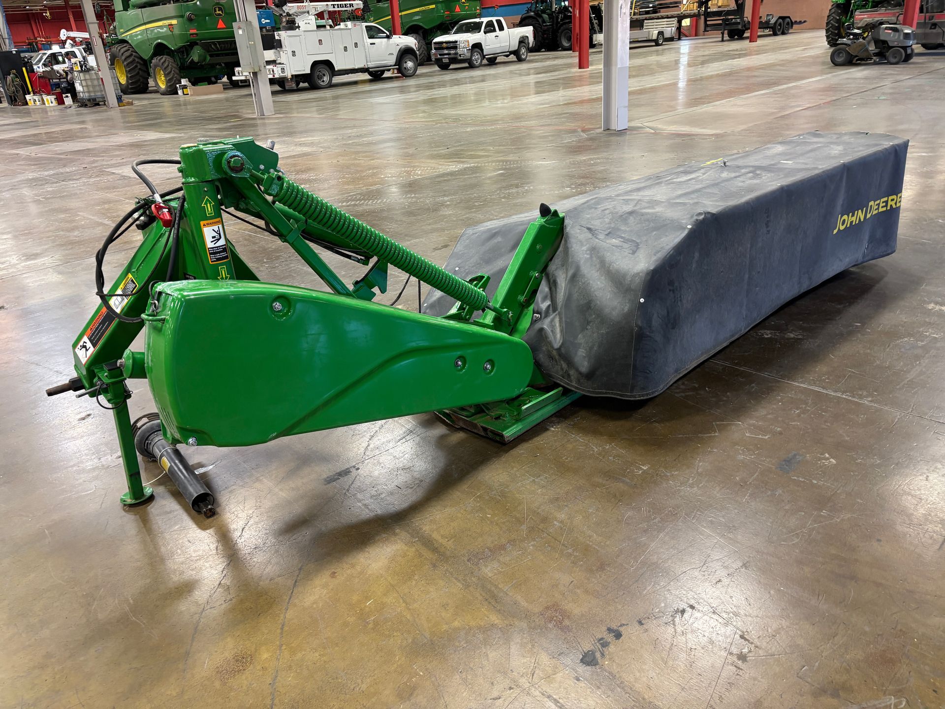 Green, covered John Deere disc mower on a concrete floor in a warehouse setting, angled shot.