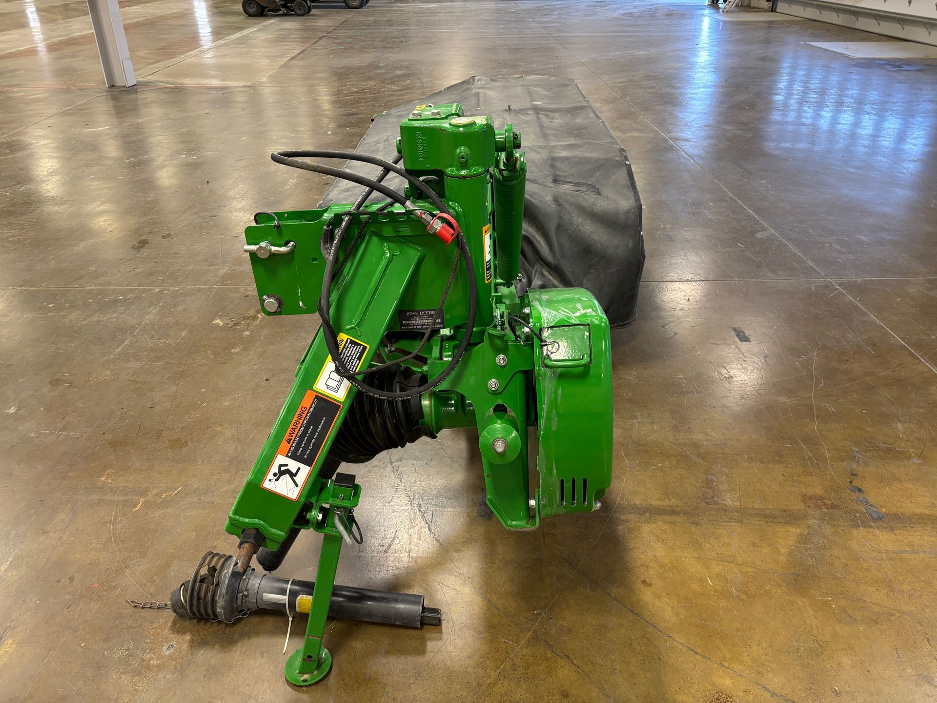 Green, covered John Deere disc mower on a concrete floor in a warehouse setting.
