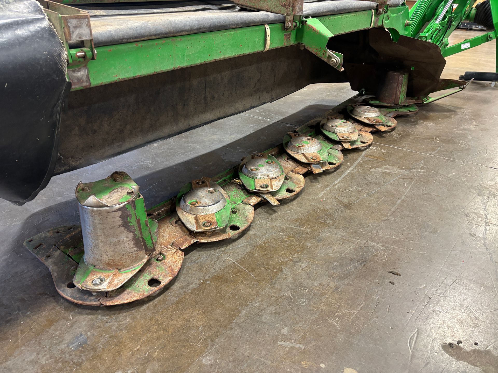 Green farm mower blades resting on a concrete floor, angled shot.