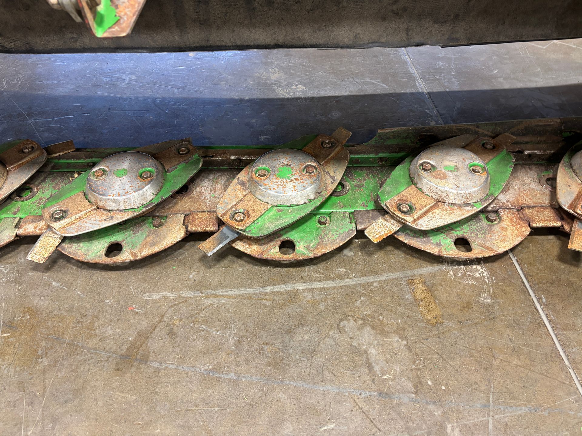 Green farm mower blades resting on a concrete floor, closeup shot.
