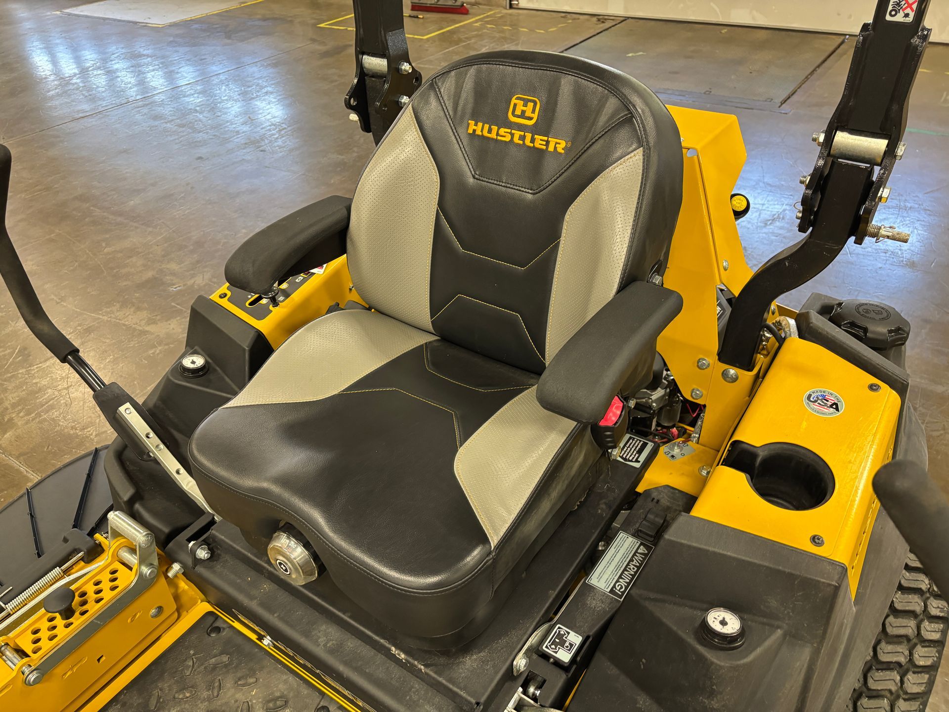 Yellow and black Hustler lawn mower seat with armrests, cup holder, and control levers.
