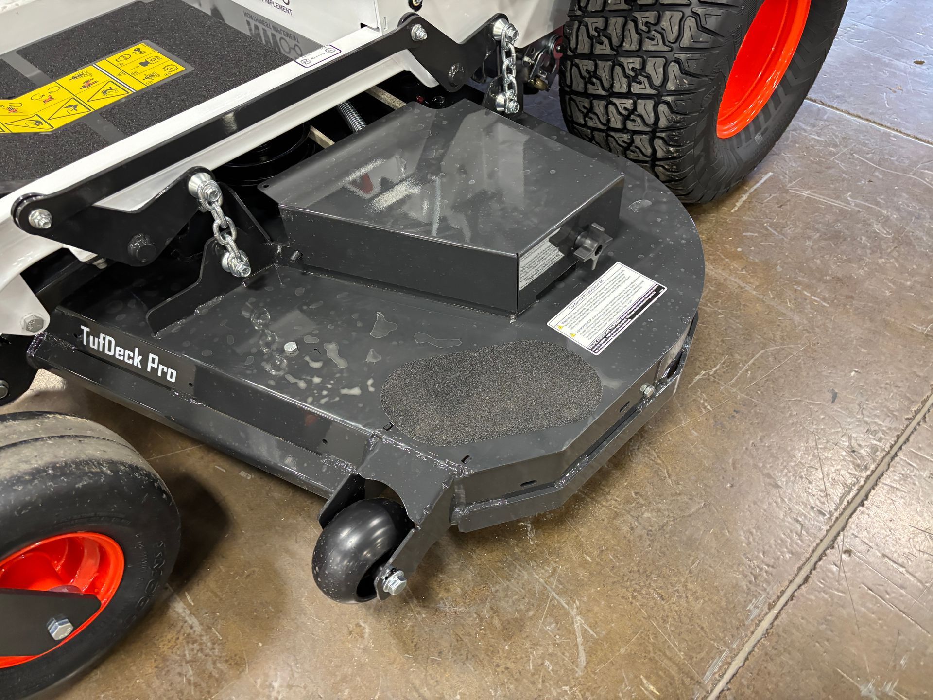 Close-up of a gray lawn mower deck with a black top on a concrete floor. The wheel is orange.