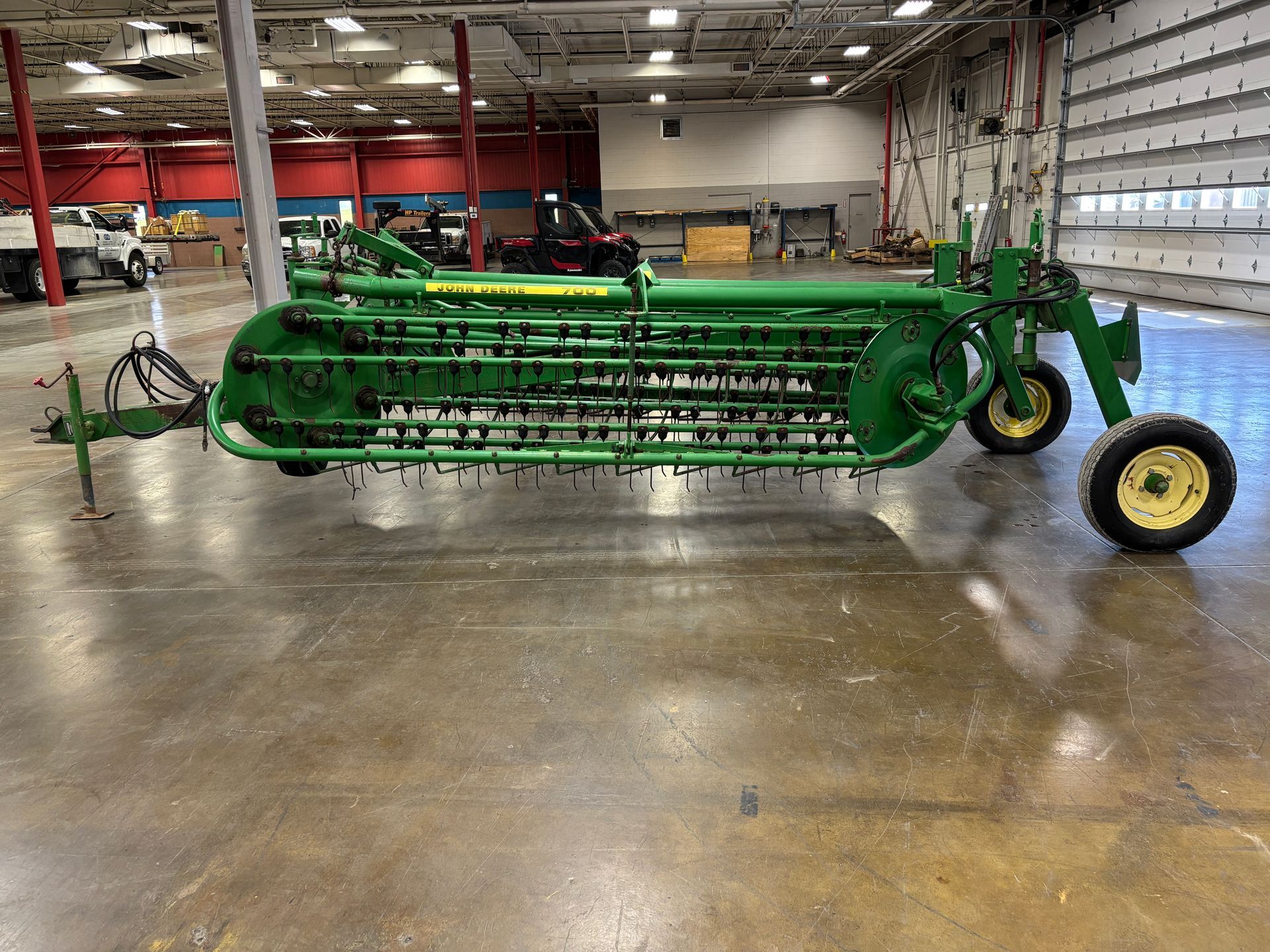 John Deere hay rake with green frame, black tines, and yellow wheels on a concrete floor.