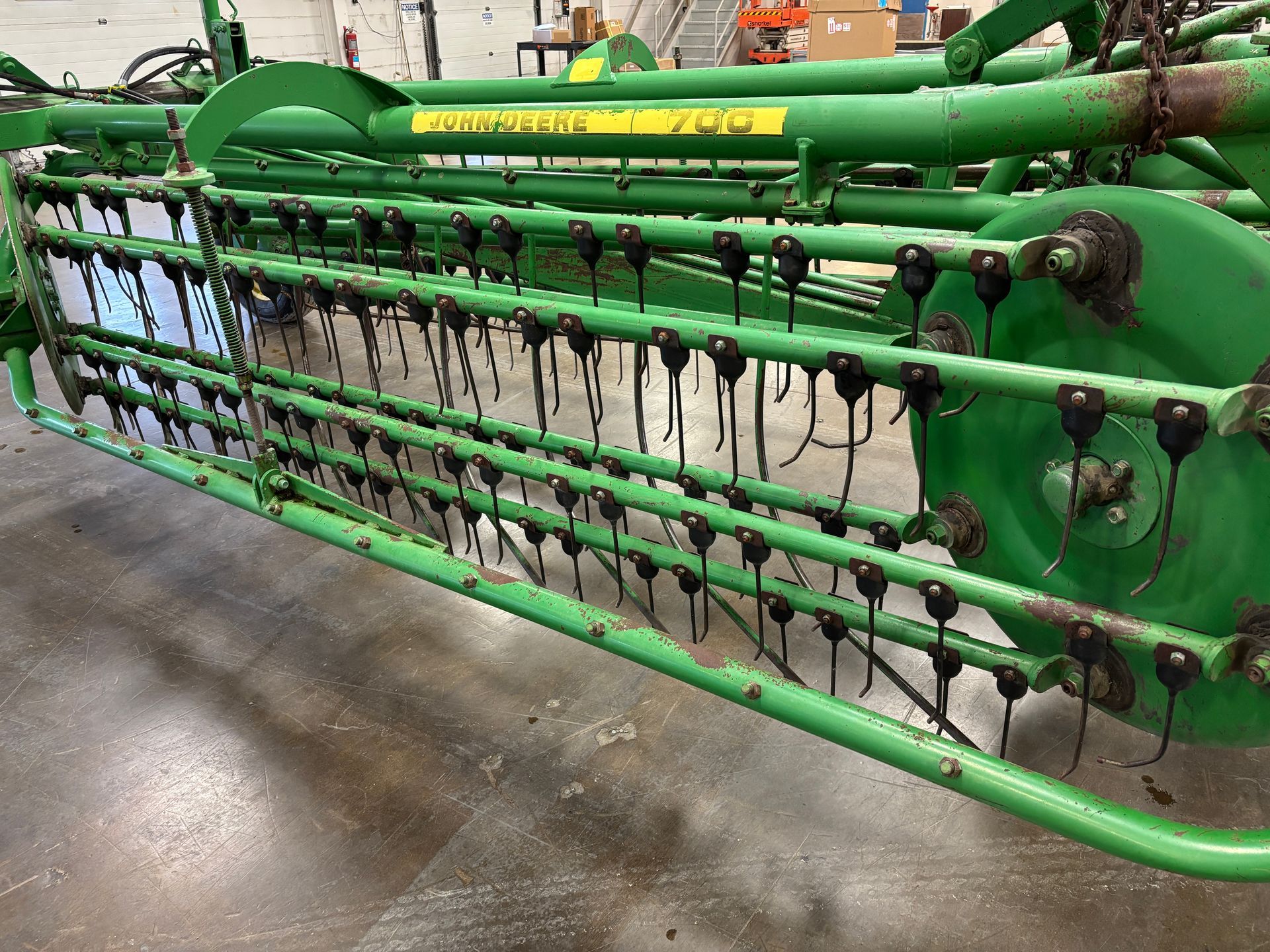 Green John Deere hay rake with curved tines, parked on a concrete floor in a workshop.