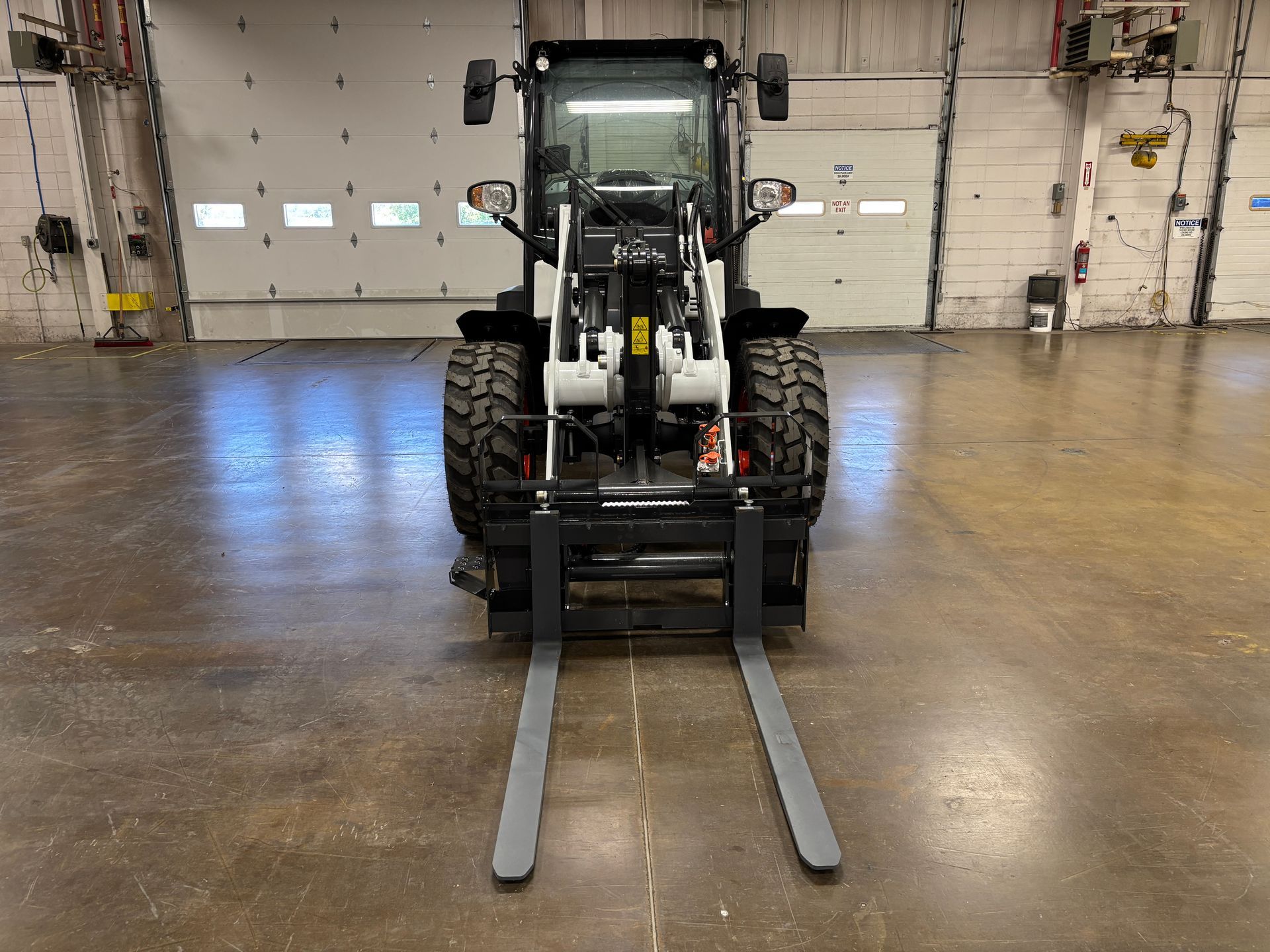 Bobcat skid-steer loader with forklift attachment, indoors. Gray forks, black machine, and concrete floor.