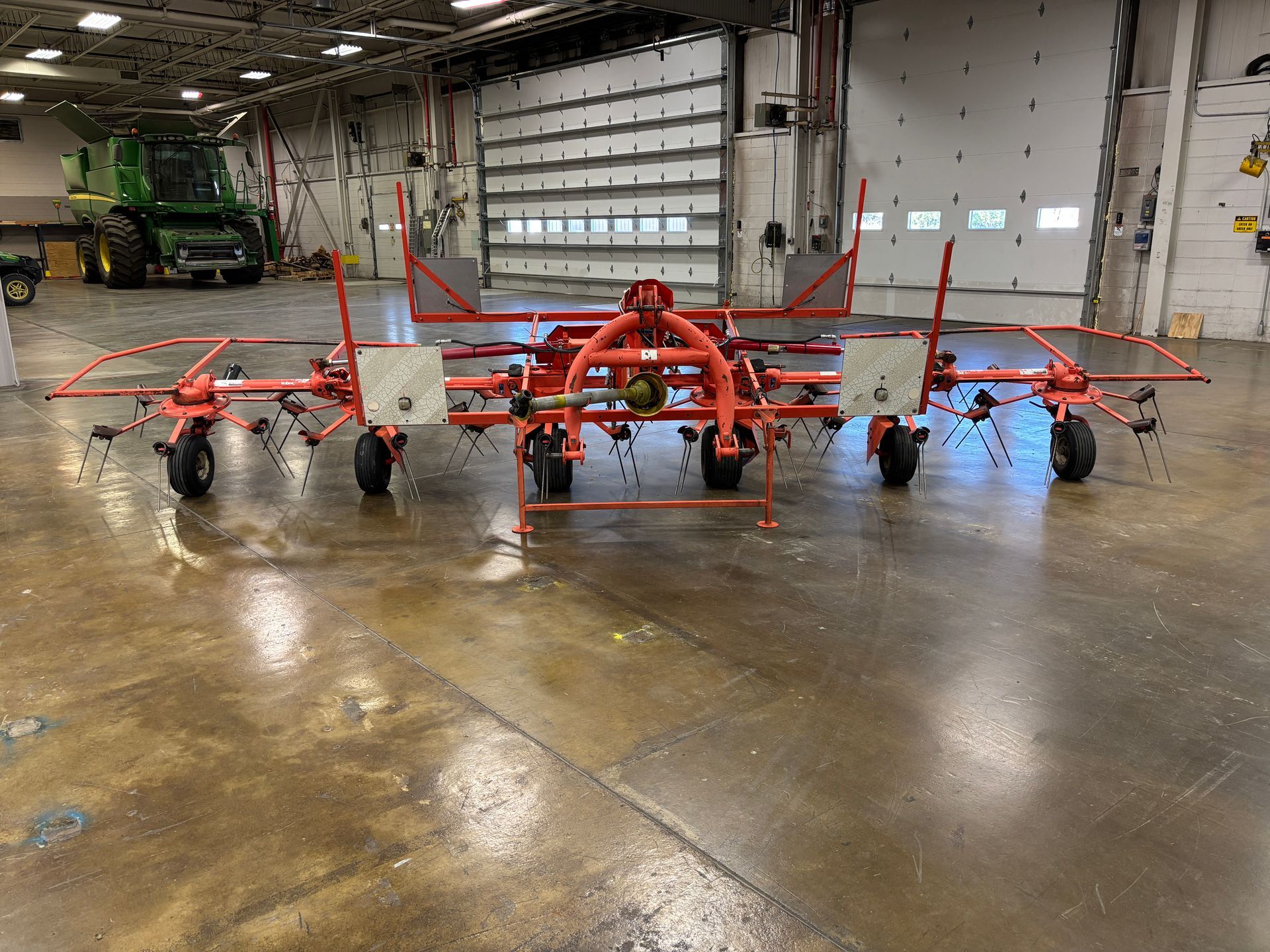 Orange hay rake on a shiny, brown floor, with a green tractor in the background. White door behind it.