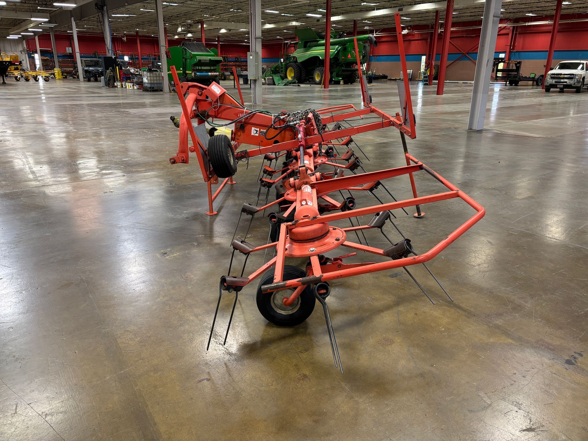 An orange hay rake on a concrete floor in a large building with other farm equipment.