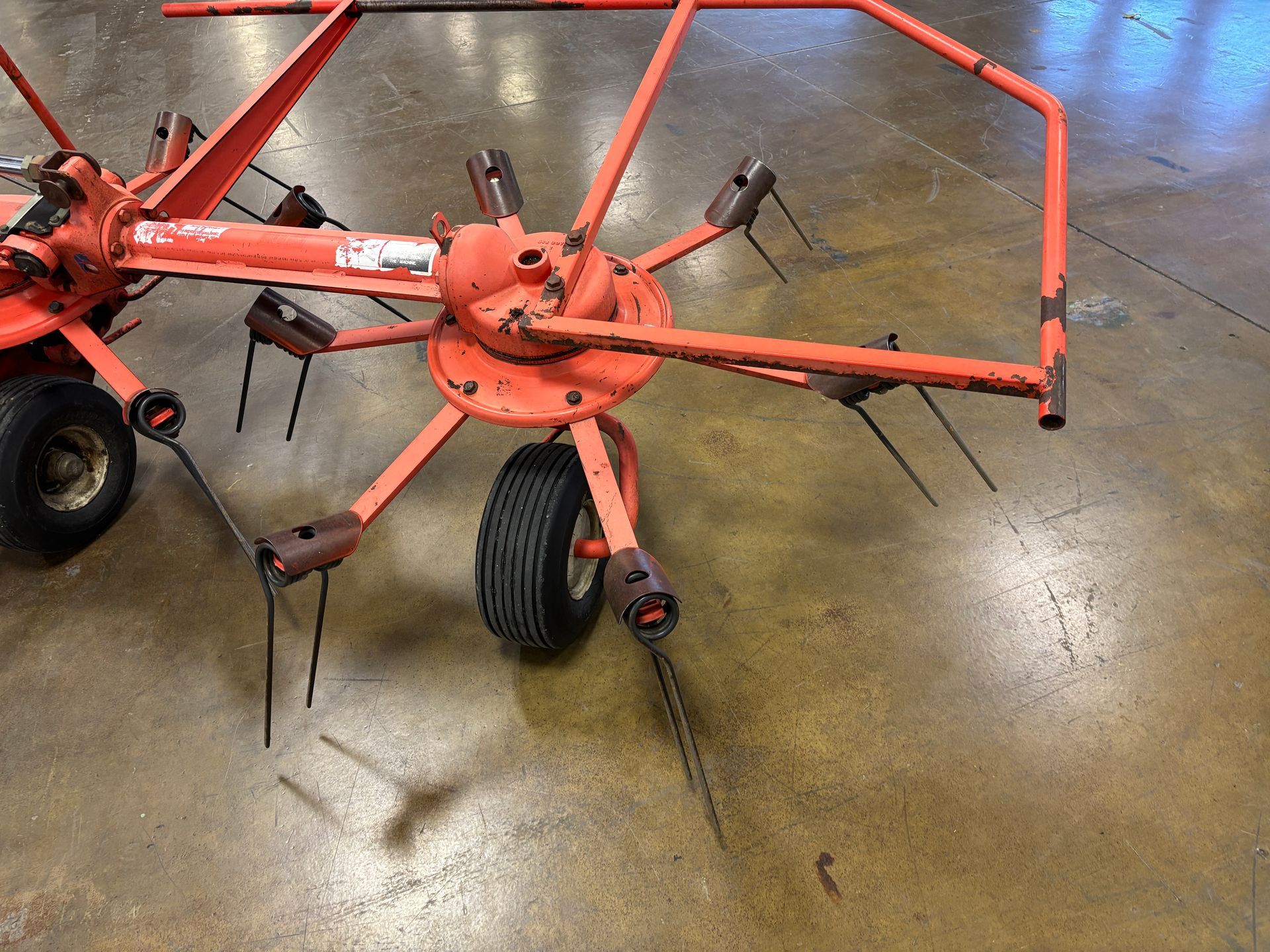 Orange hay rake on a concrete floor with black tires and metal tines.