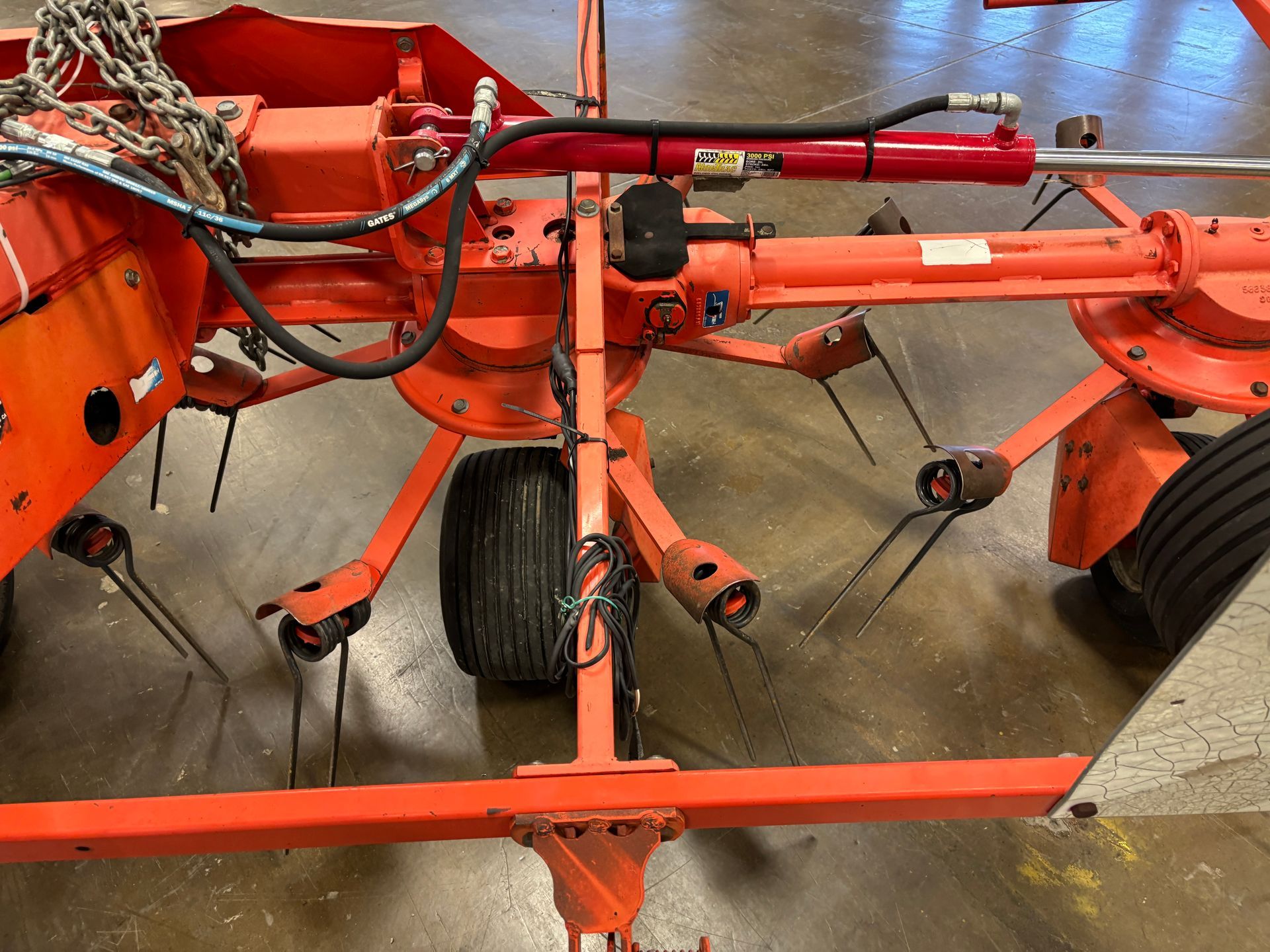 Orange hay rake with black tines, wheels, and a hydraulic cylinder in a workshop.