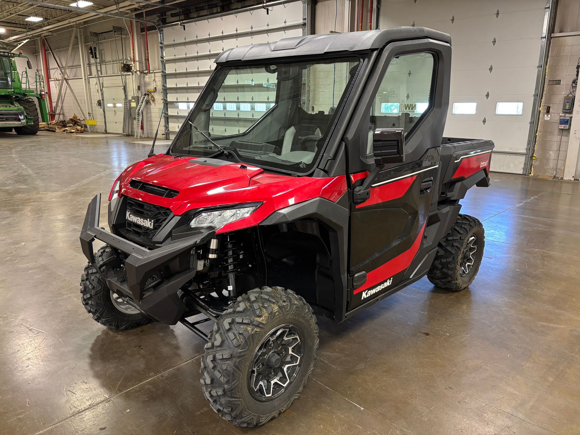 Red and black Kawasaki side-by-side vehicle with a windshield and cabin, parked indoors.