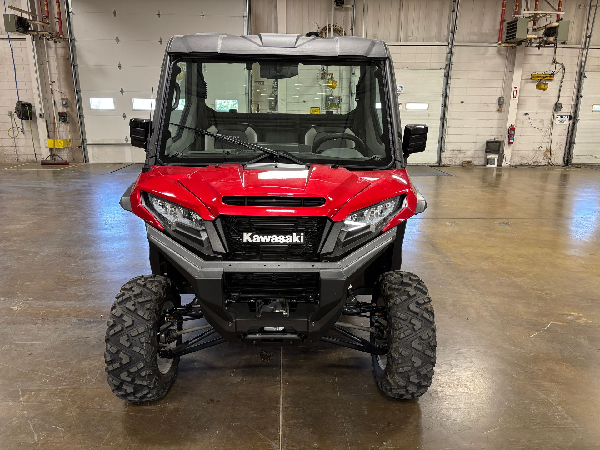 Red Kawasaki UTV parked indoors, facing the viewer, large tires, black front bumper, and glass windshield.