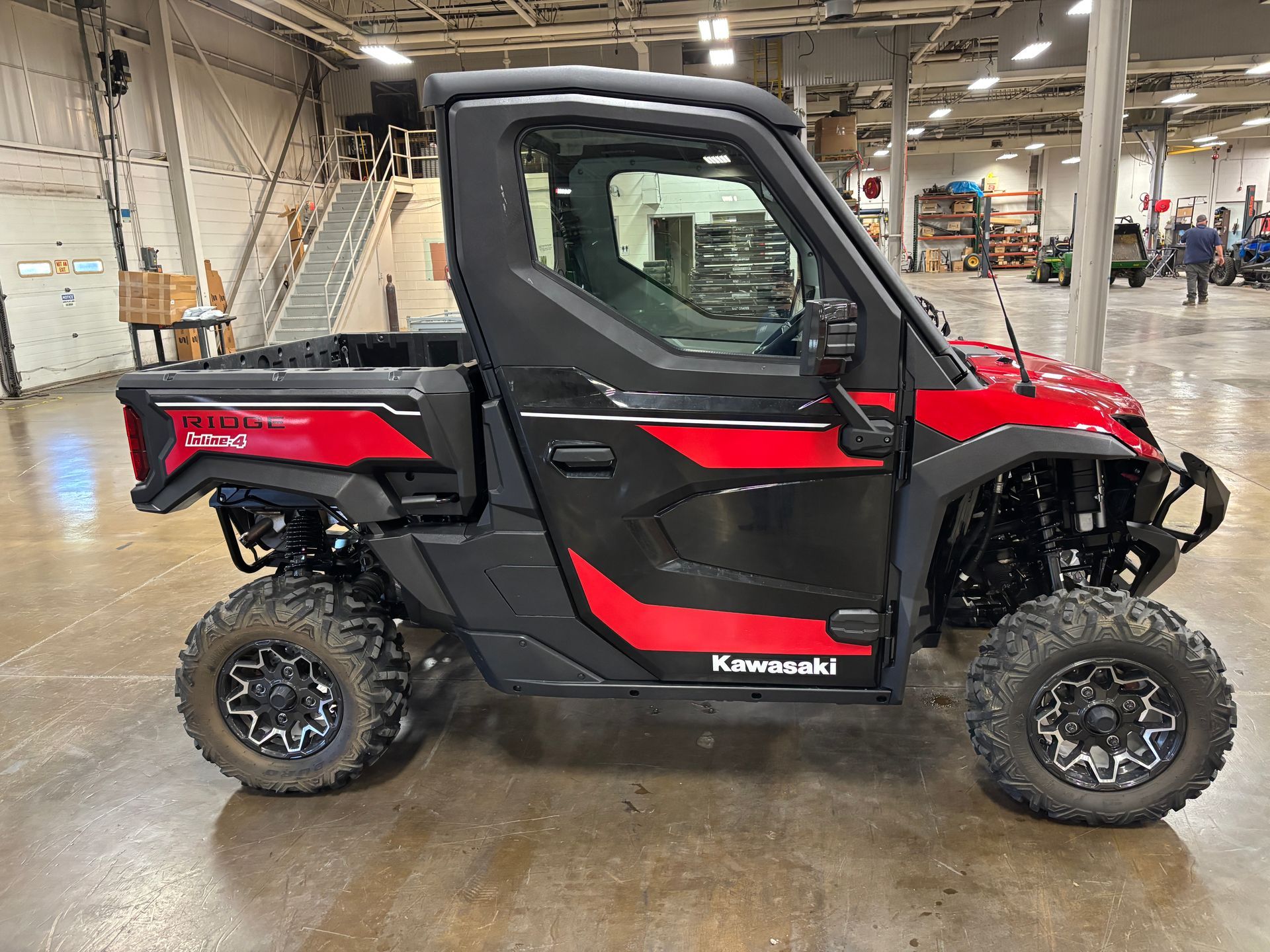 Red and black Kawasaki UTV with black wheels and a cab, parked indoors.