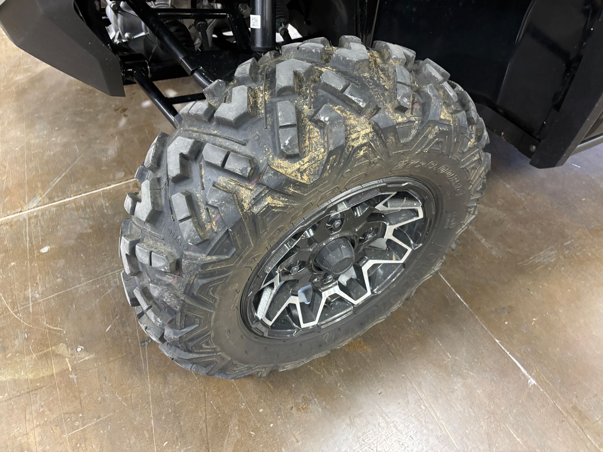 Black off-road tire on a side-by-side vehicle, covered in dirt, sitting on a concrete surface.