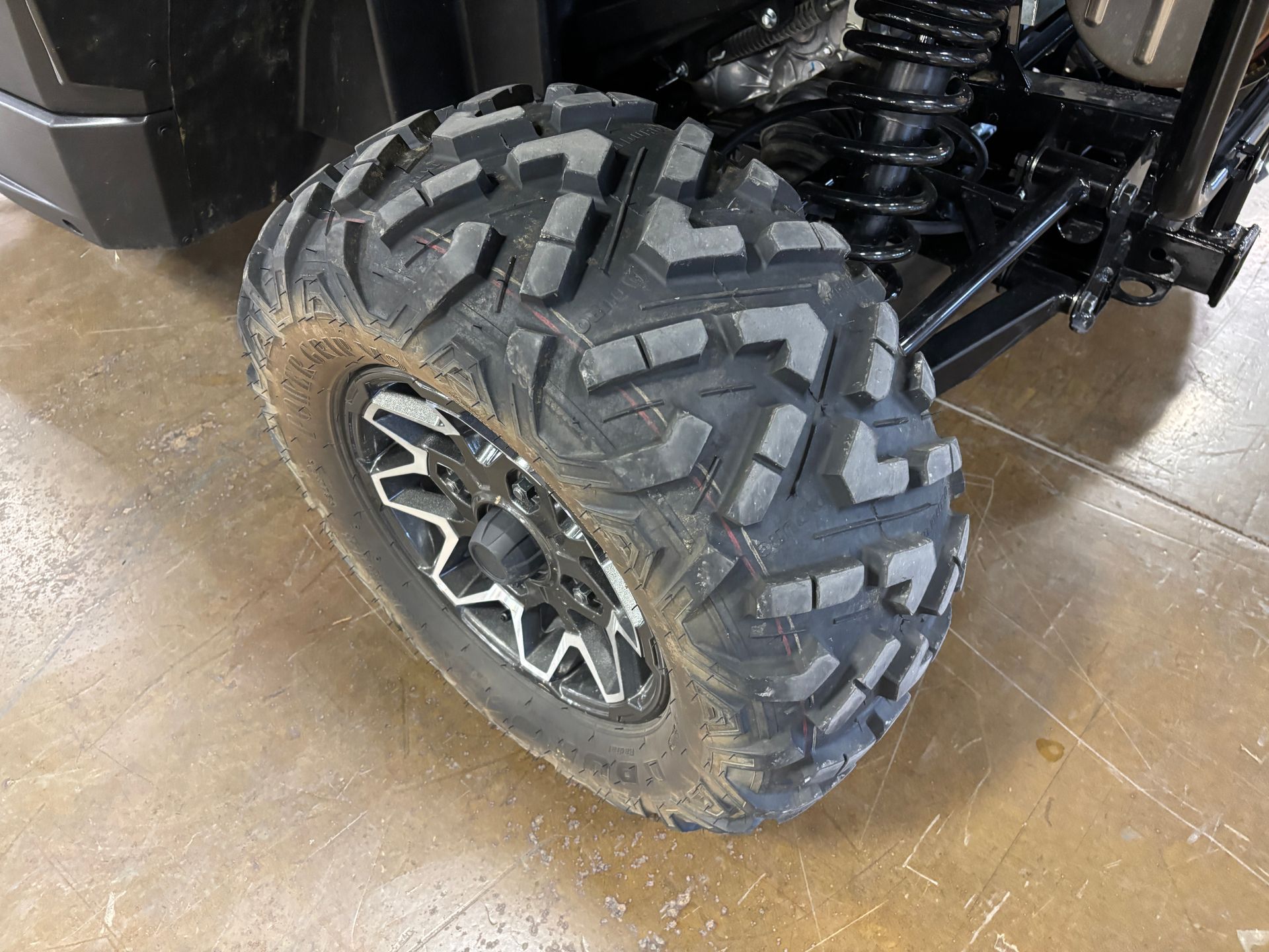 Close-up of a black tire with a complex tread pattern on a silver and black wheel, on a dusty surface.