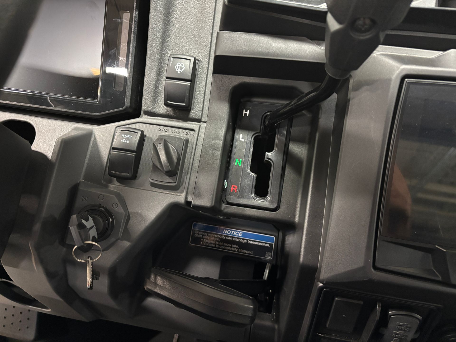 Close-up of a side-by-side vehicle's dashboard, showing a gear selector, various switches, a key, and two screens.