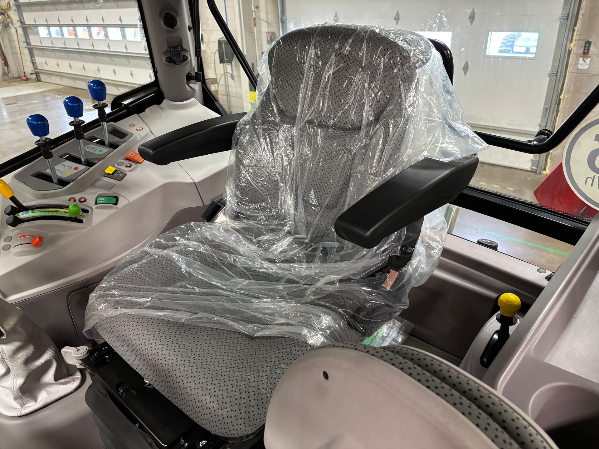 Interior view of a tractor cab. Seat is covered in plastic. Levers, controls, and a window are visible.