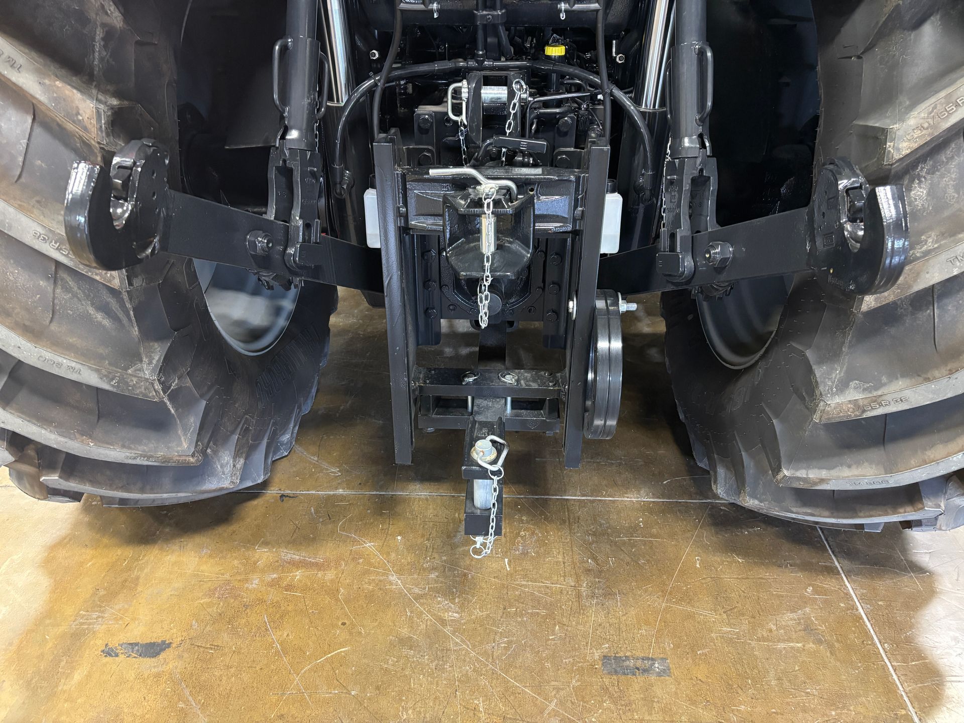 Rear view of a black tractor with large tires and a towing hitch, sitting on a concrete floor.