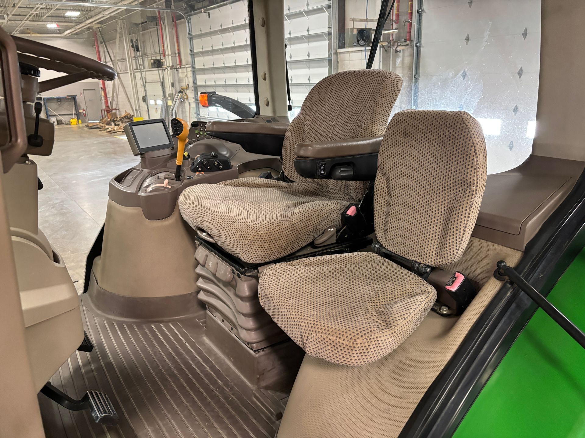 Interior view of a tractor cab with tan patterned seats, control panel, and flooring; green side panel.