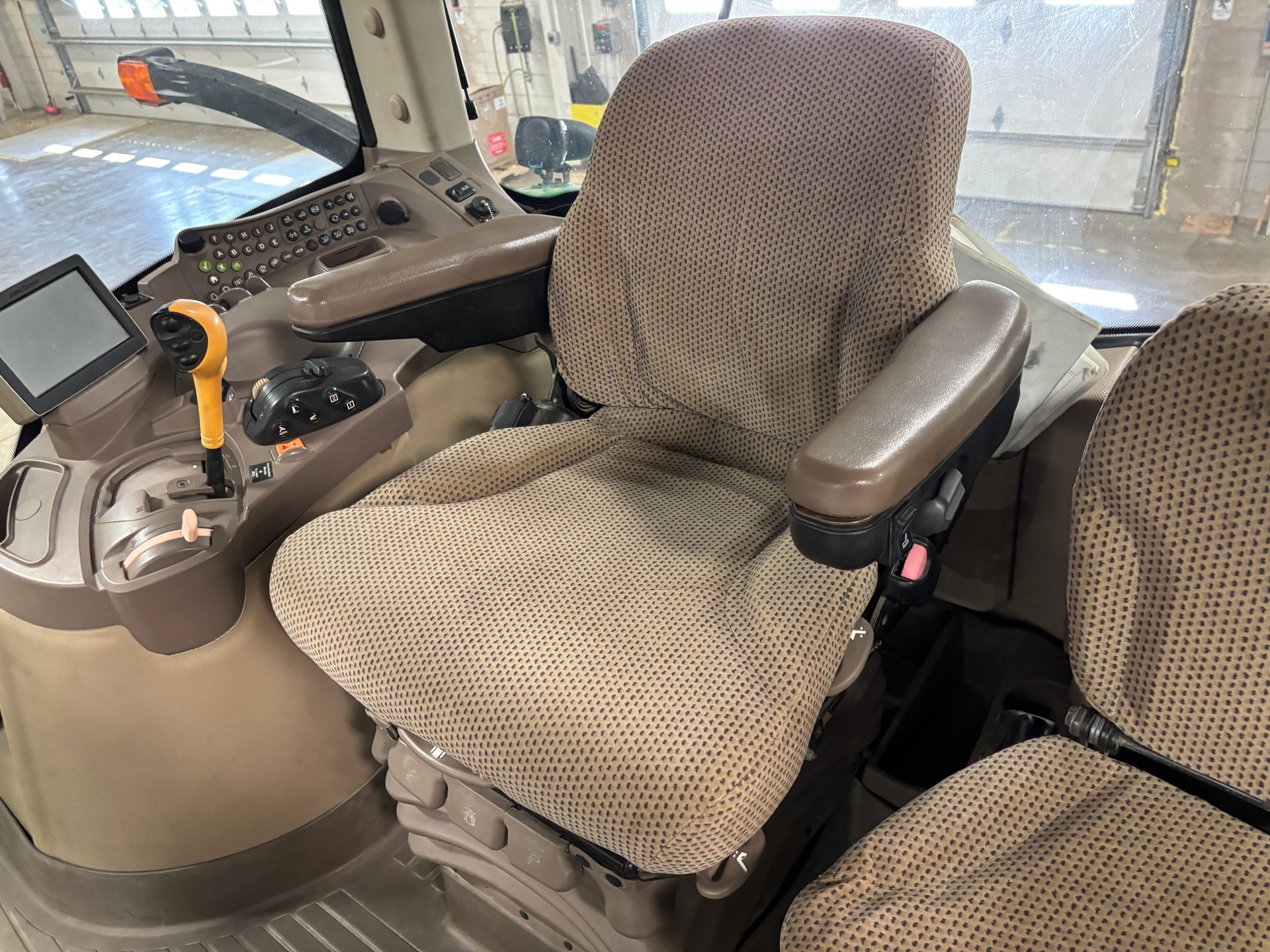 Interior of a tractor cab showing the driver's seat and controls. Brown fabric seat, gray controls, and a display screen.