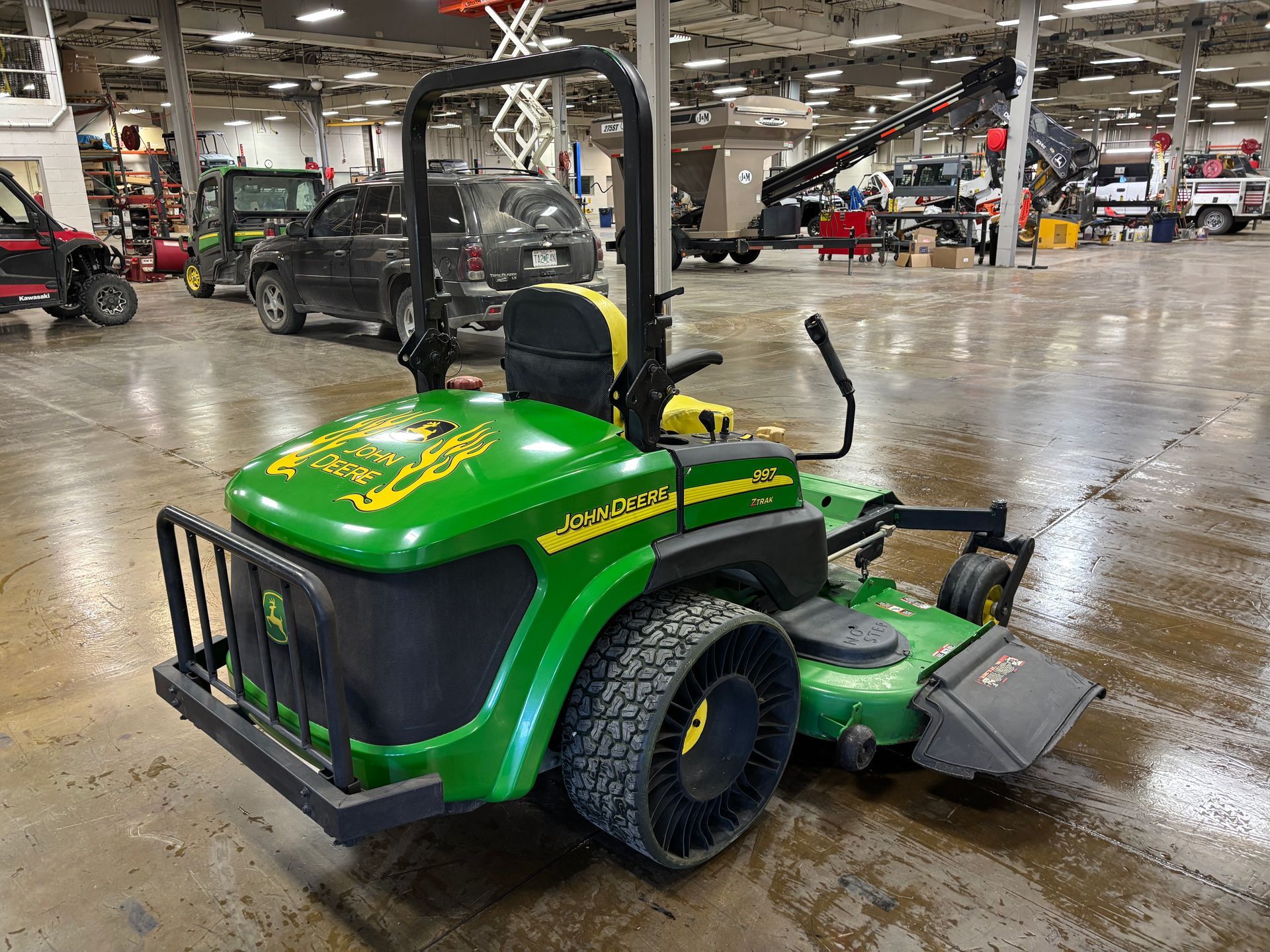 John Deere zero-turn lawnmower with green and yellow accents, large tires, and roll bar inside a large warehouse.
