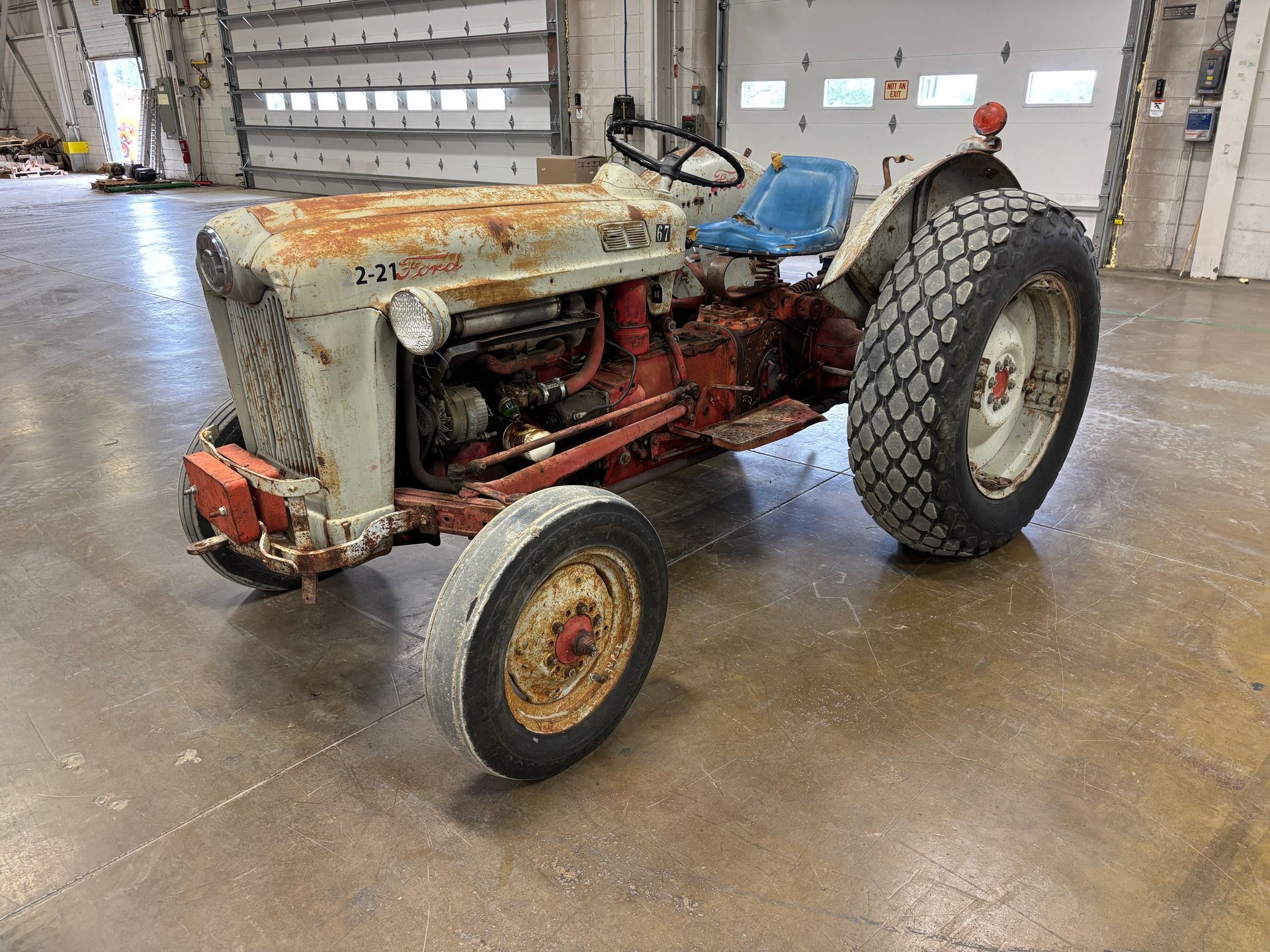 Rusty, vintage Ford tractor in a warehouse. Gray and red paint, blue seat, large tires.