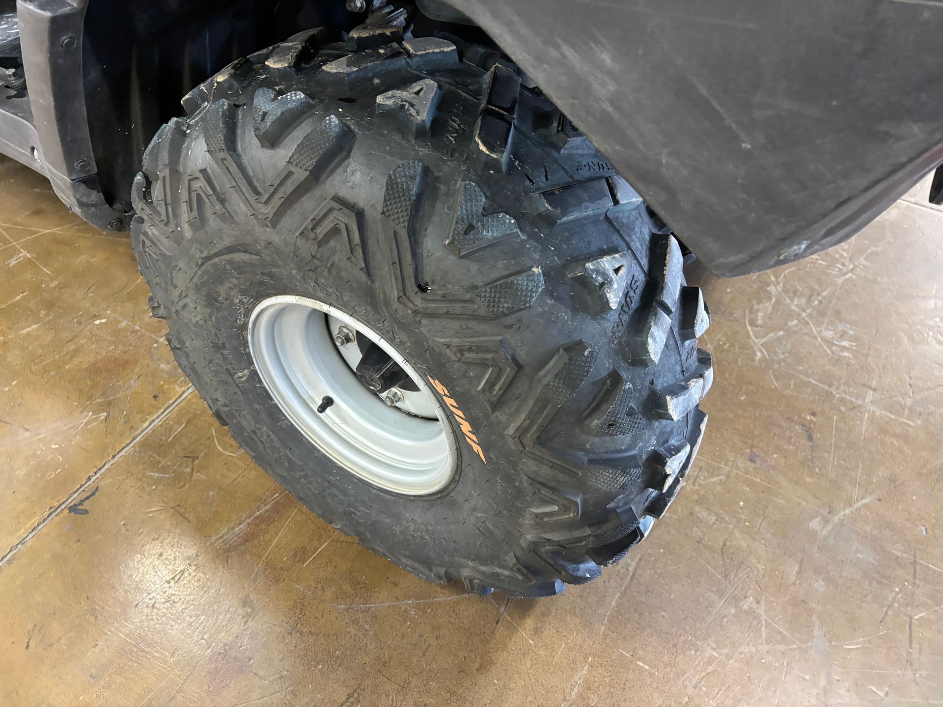Close-up of an ATV tire. Black tire with deep tread, mounted on a silver rim. Brown floor visible.