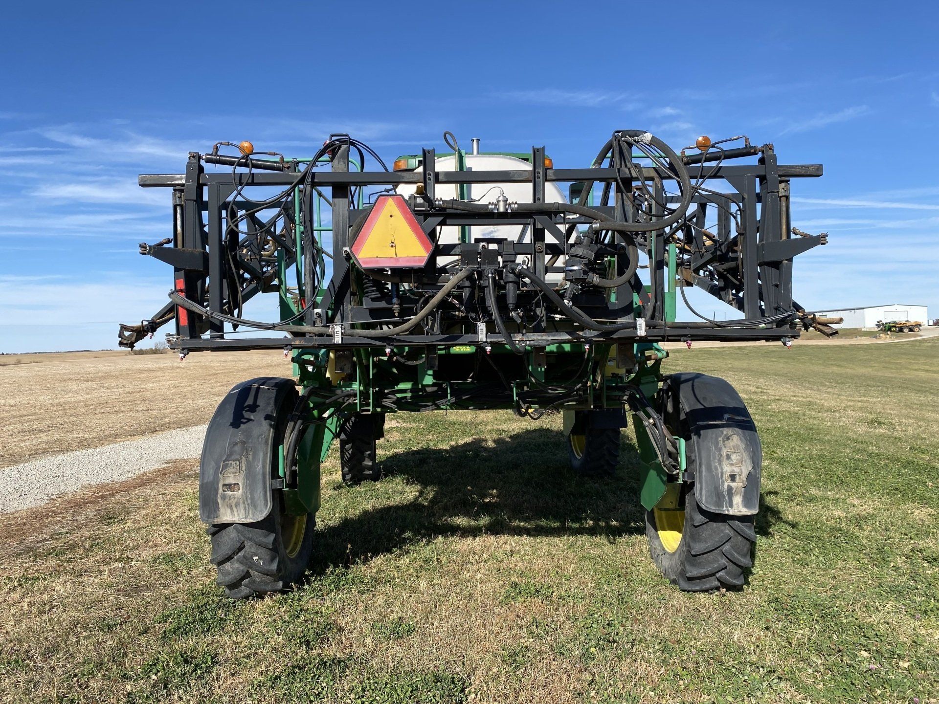 John Deere agricultural sprayer on a field, facing the camera. Green and black with a white tank and yellow safety triangle.