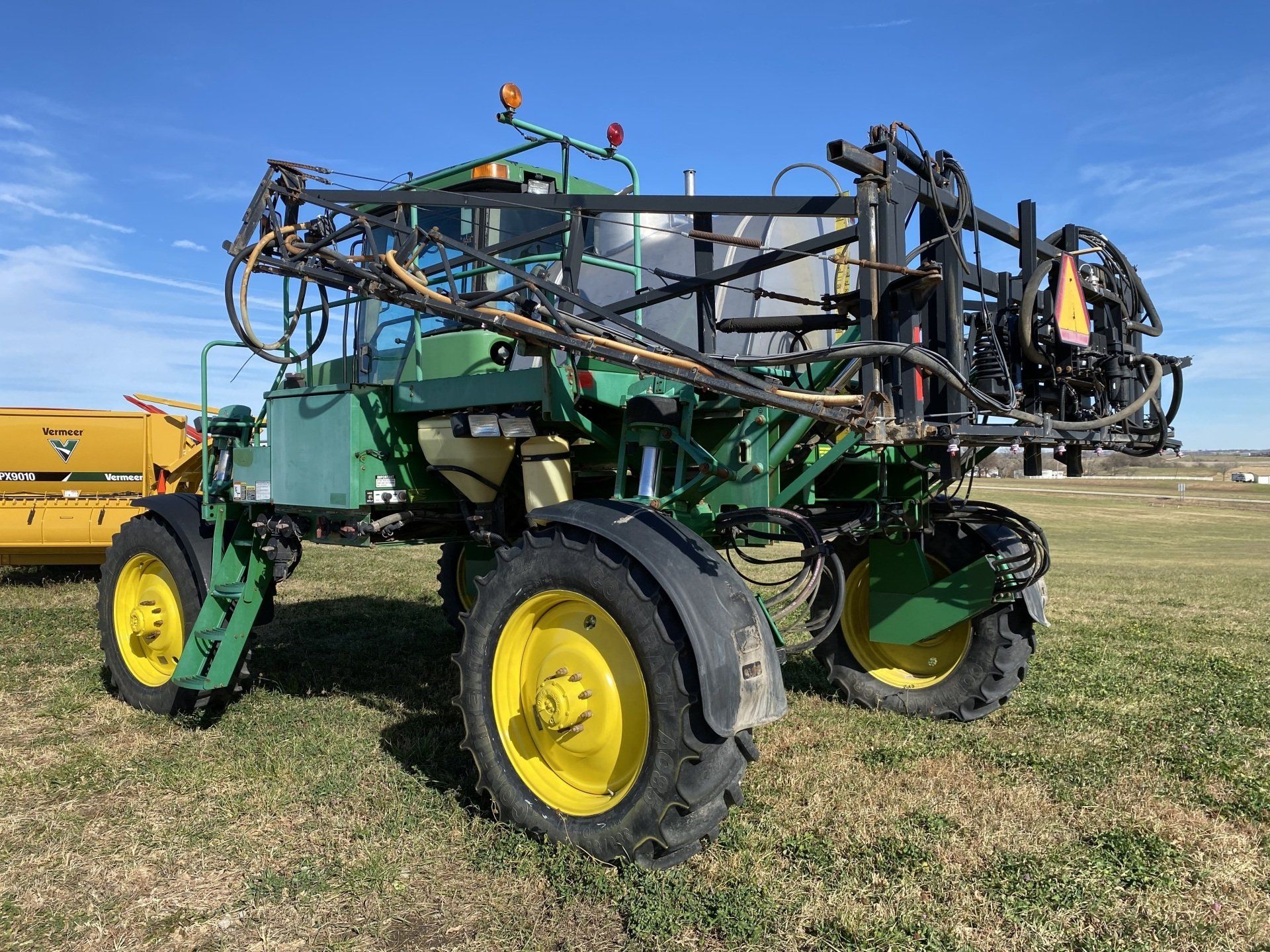 Green John Deere agricultural sprayer with yellow wheels on a field under a blue sky.