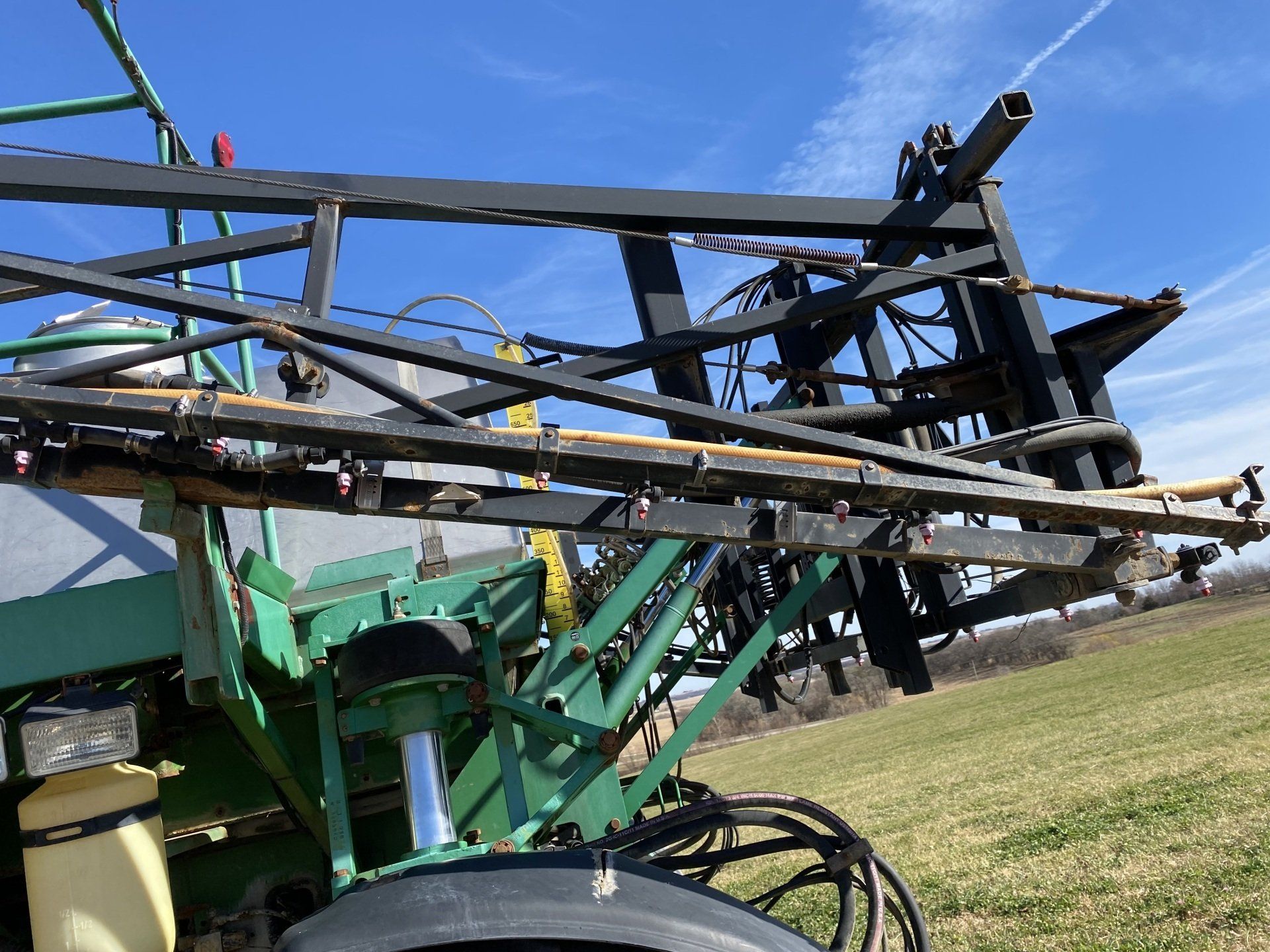 Green and black farm sprayer boom with nozzles against a blue sky.