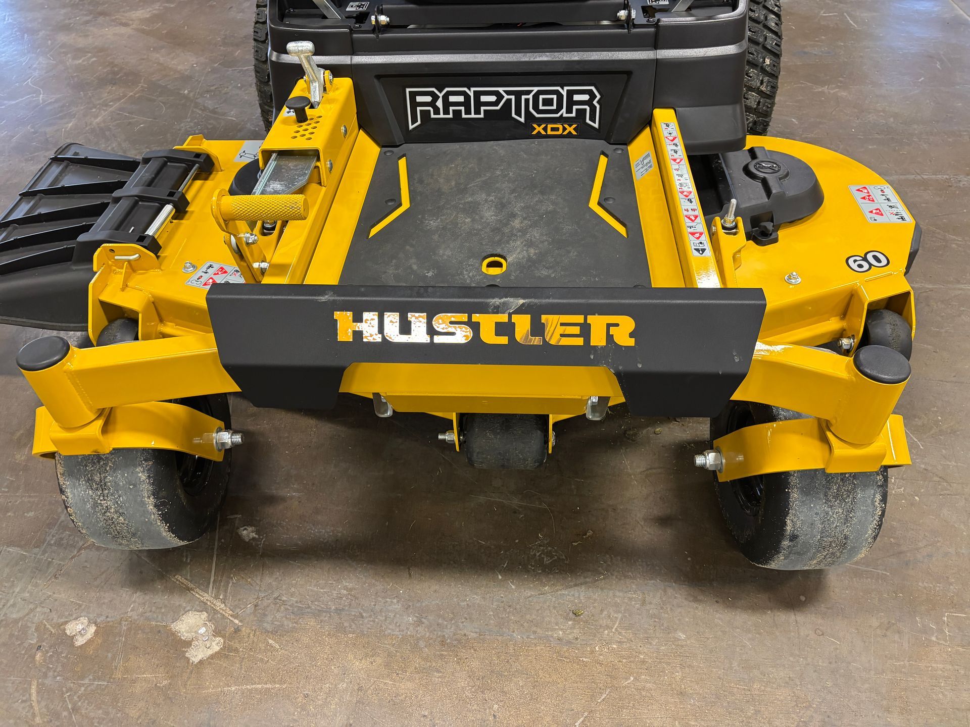 Yellow and black Hustler Raptor zero-turn mower with black deck and tires; parked on floor.