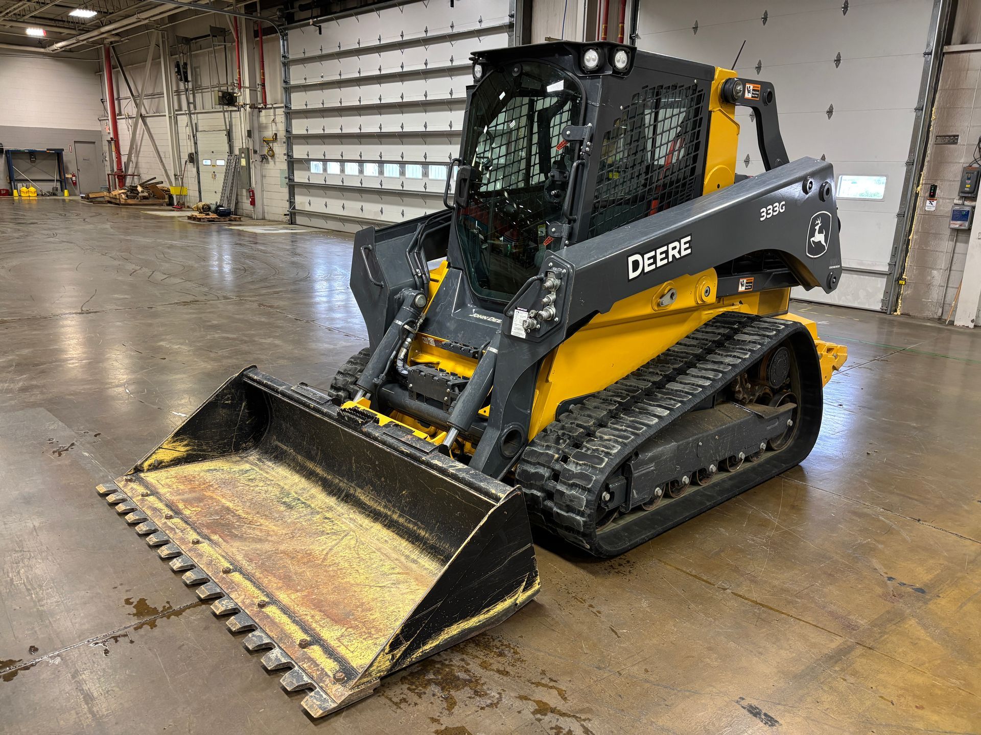 Yellow and black John Deere compact track loader with a front bucket on a gray concrete floor, angled shot.