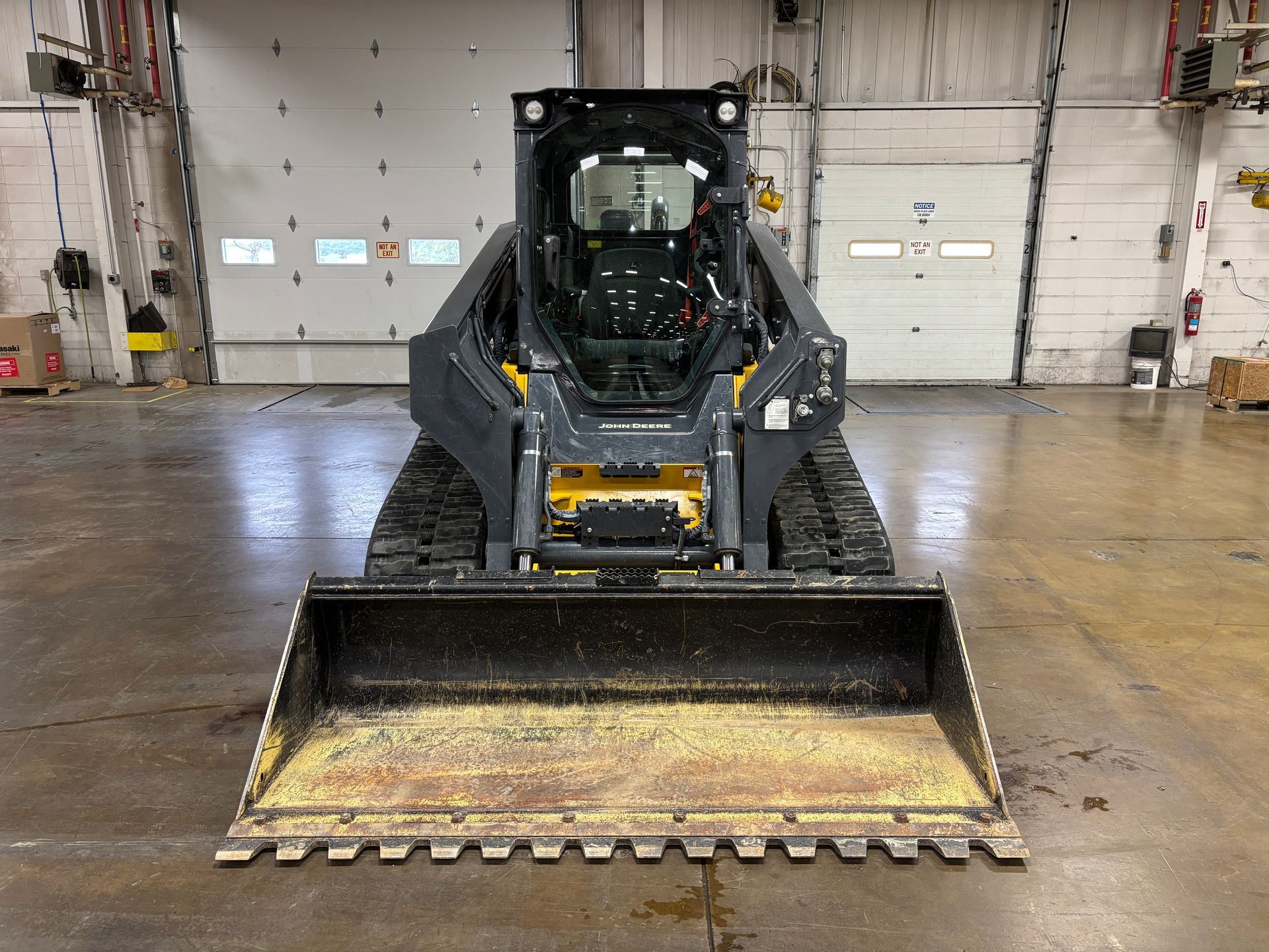 Yellow and black John Deere compact track loader with a front bucket on a gray concrete floor, front shot.