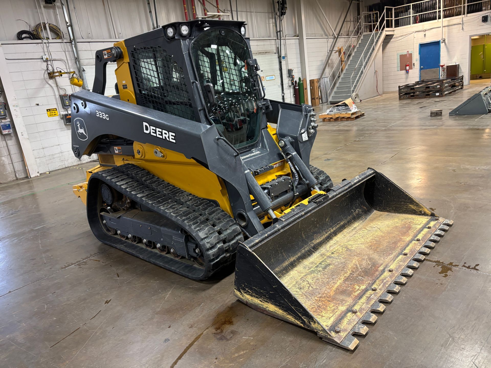 Yellow and black John Deere compact track loader with a front bucket on a gray concrete floor, front angled shot.