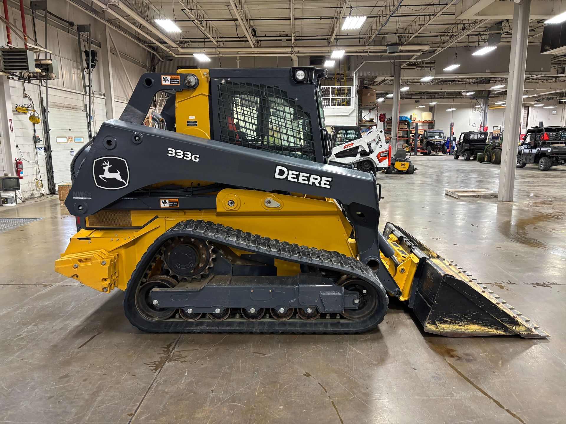 Yellow and black John Deere compact track loader with a front bucket on a gray concrete floor, side view shot.