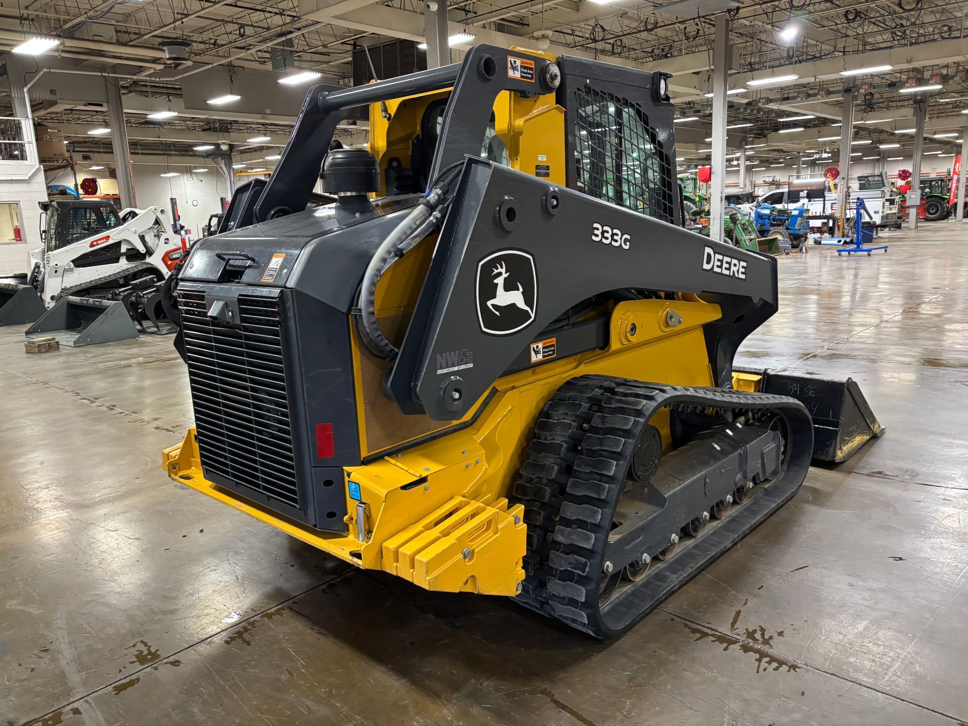 Yellow and black John Deere compact track loader with a front bucket on a gray concrete floor, back angled shot.