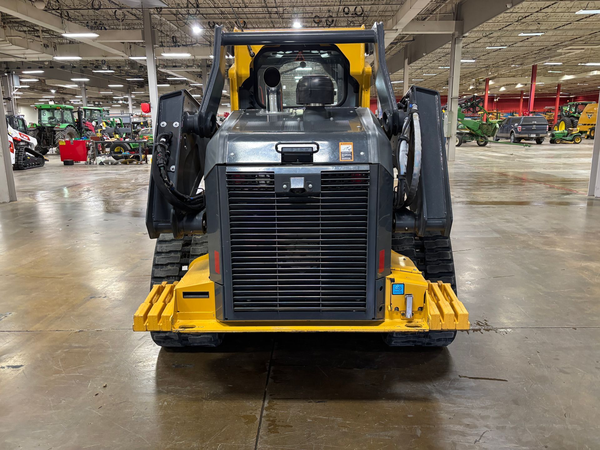 Yellow and black John Deere compact track loader with a front bucket on a gray concrete floor, back shot.