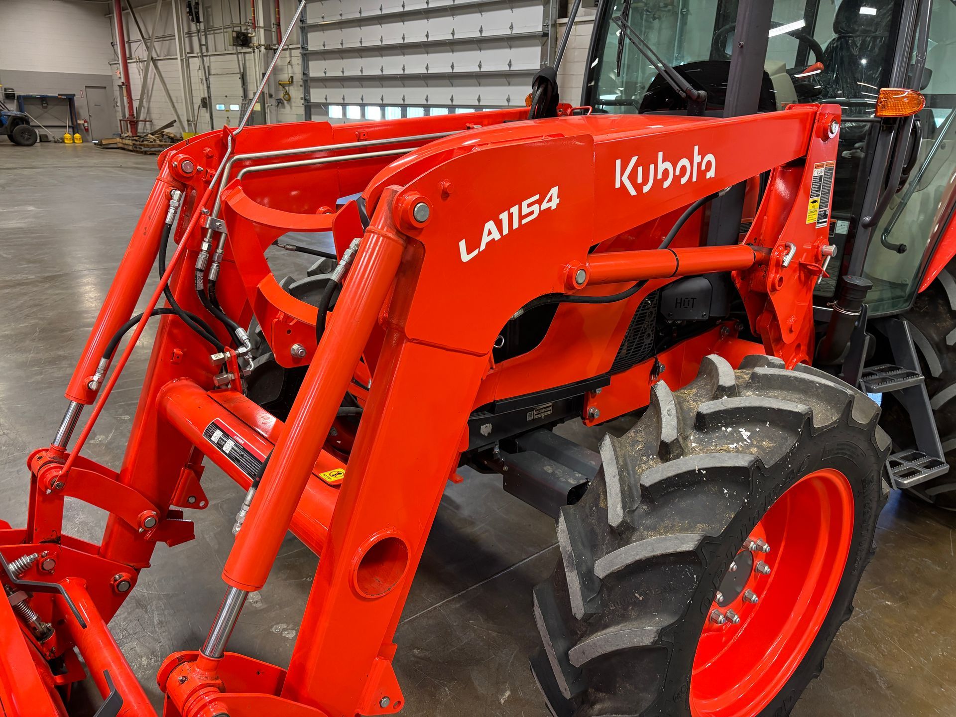 Kubota LA1154 front-end loader attached to a tractor, bright orange, parked indoors, viewed from the side.