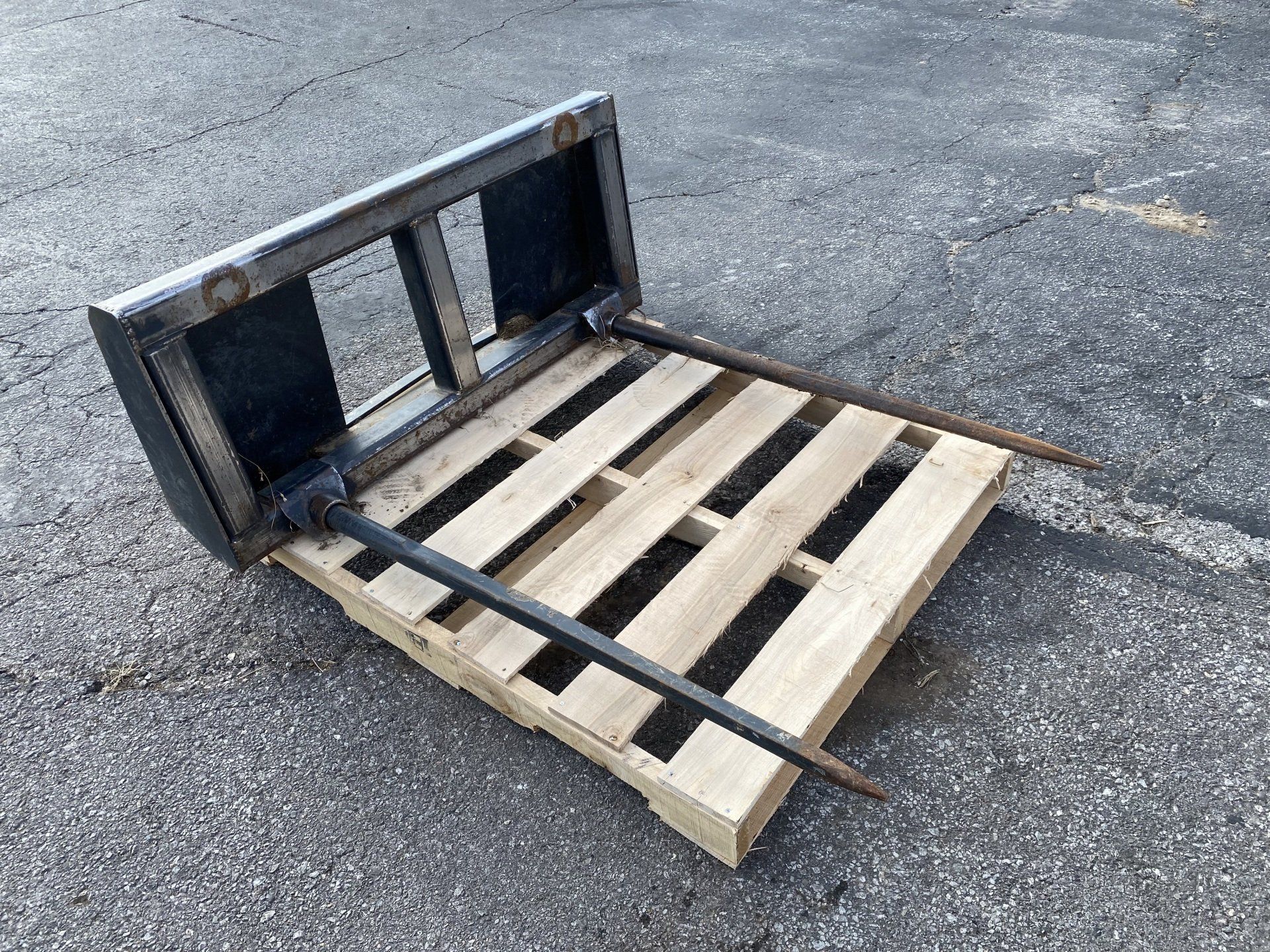 Forklift pallet forks on a wooden pallet, resting on a paved surface.