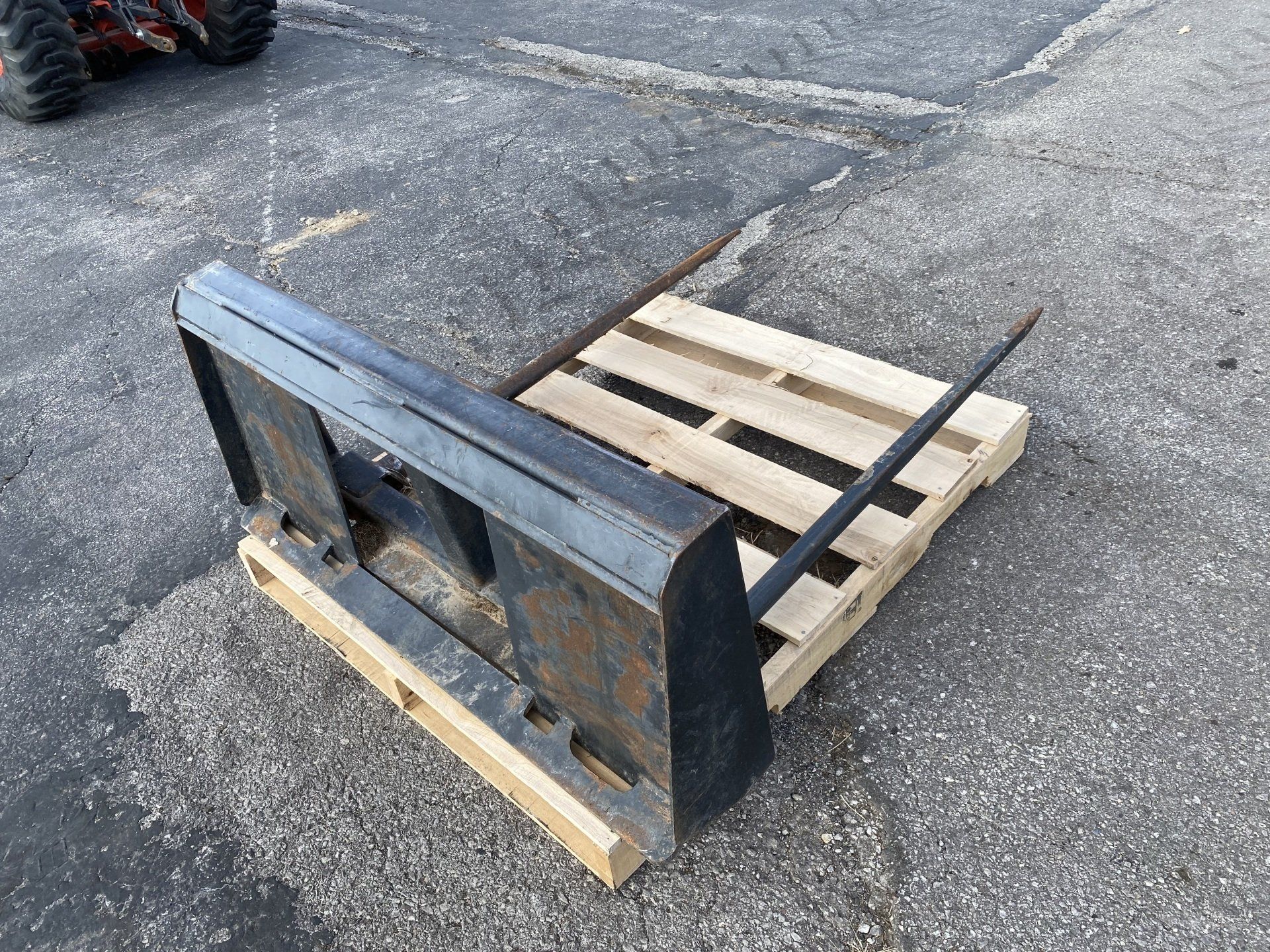 Forklift attachment on a wooden pallet, resting on pavement. Black metal with a single visible tine.