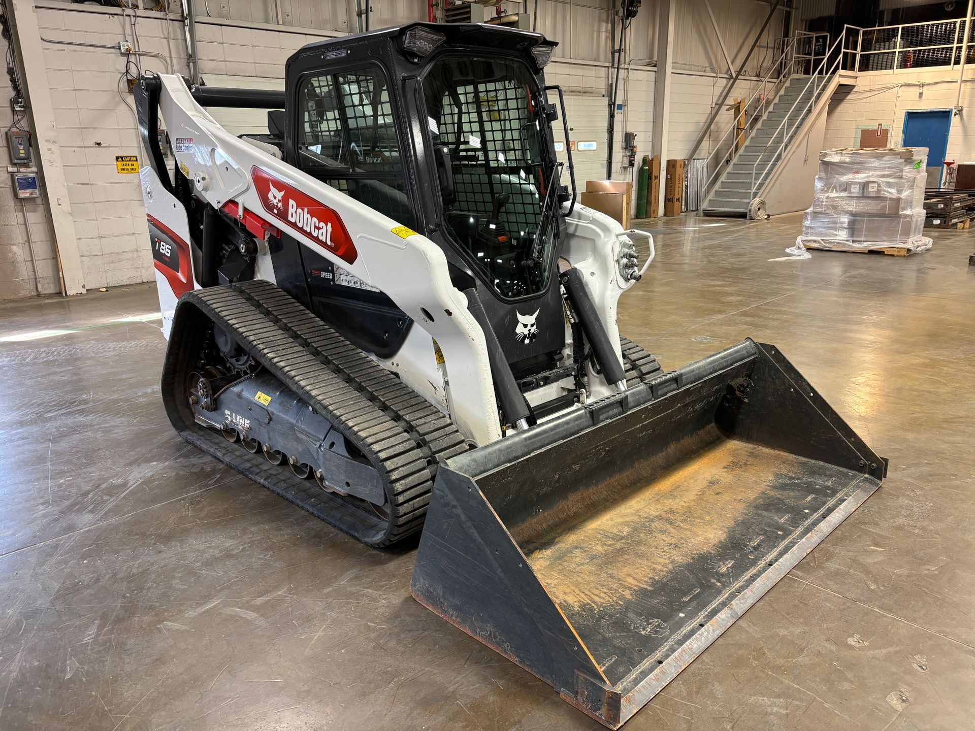White Bobcat compact track loader with bucket in a warehouse, front angled shot.