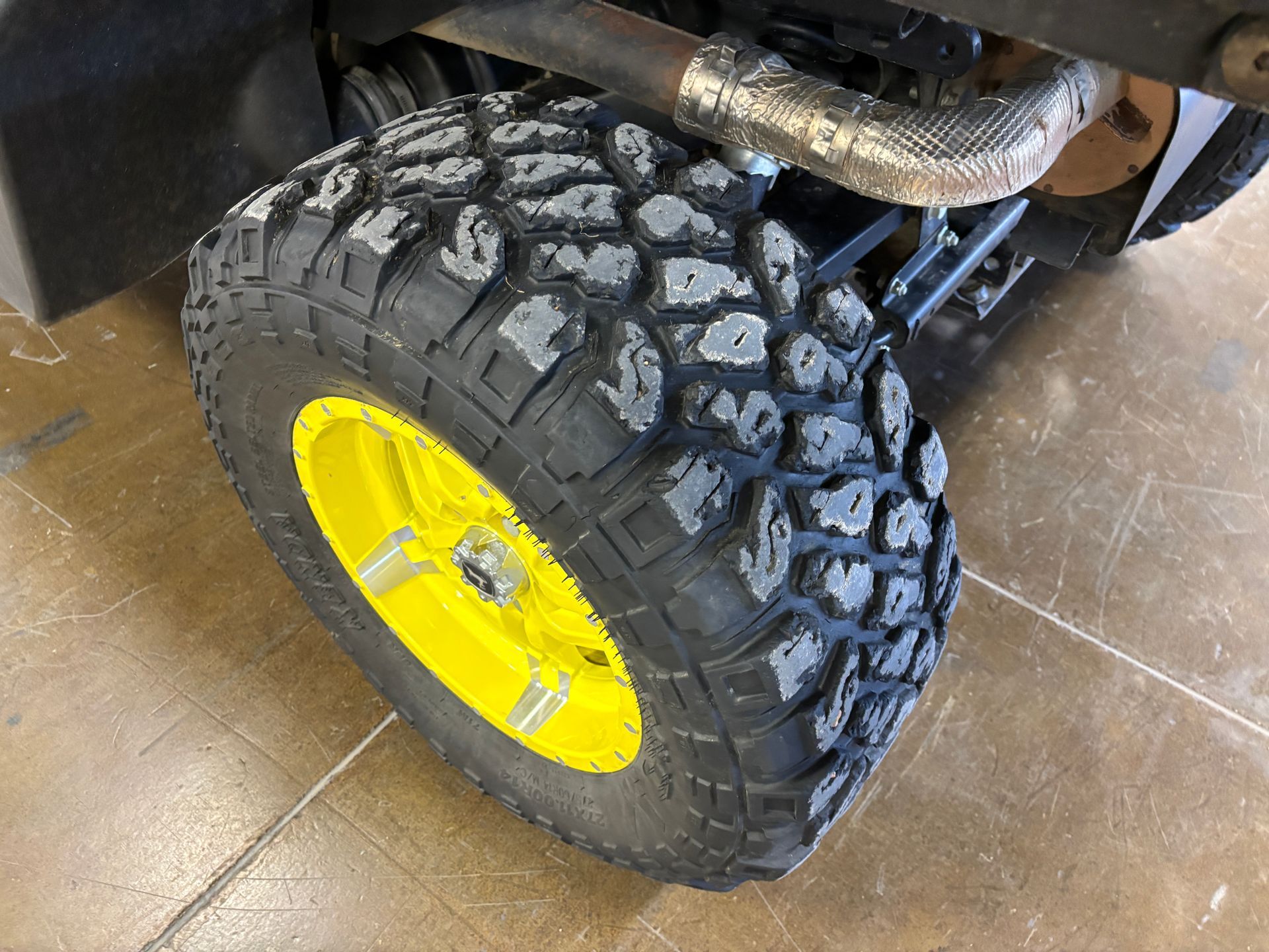 Close-up of a black off-road tire on a yellow rim. A vehicle's suspension and exhaust are visible.