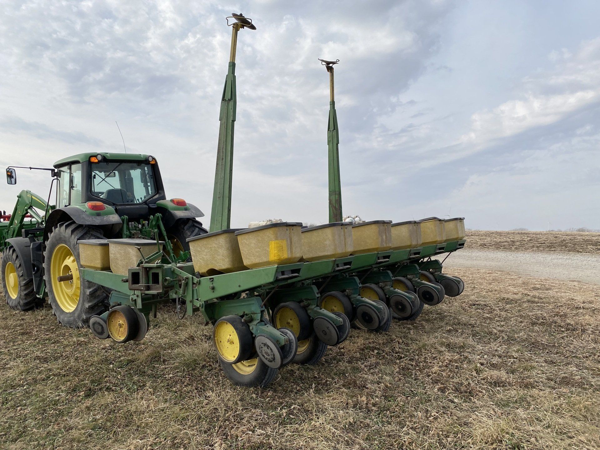 Green tractor pulling a planter in a field, planting seeds under a cloudy sky.
