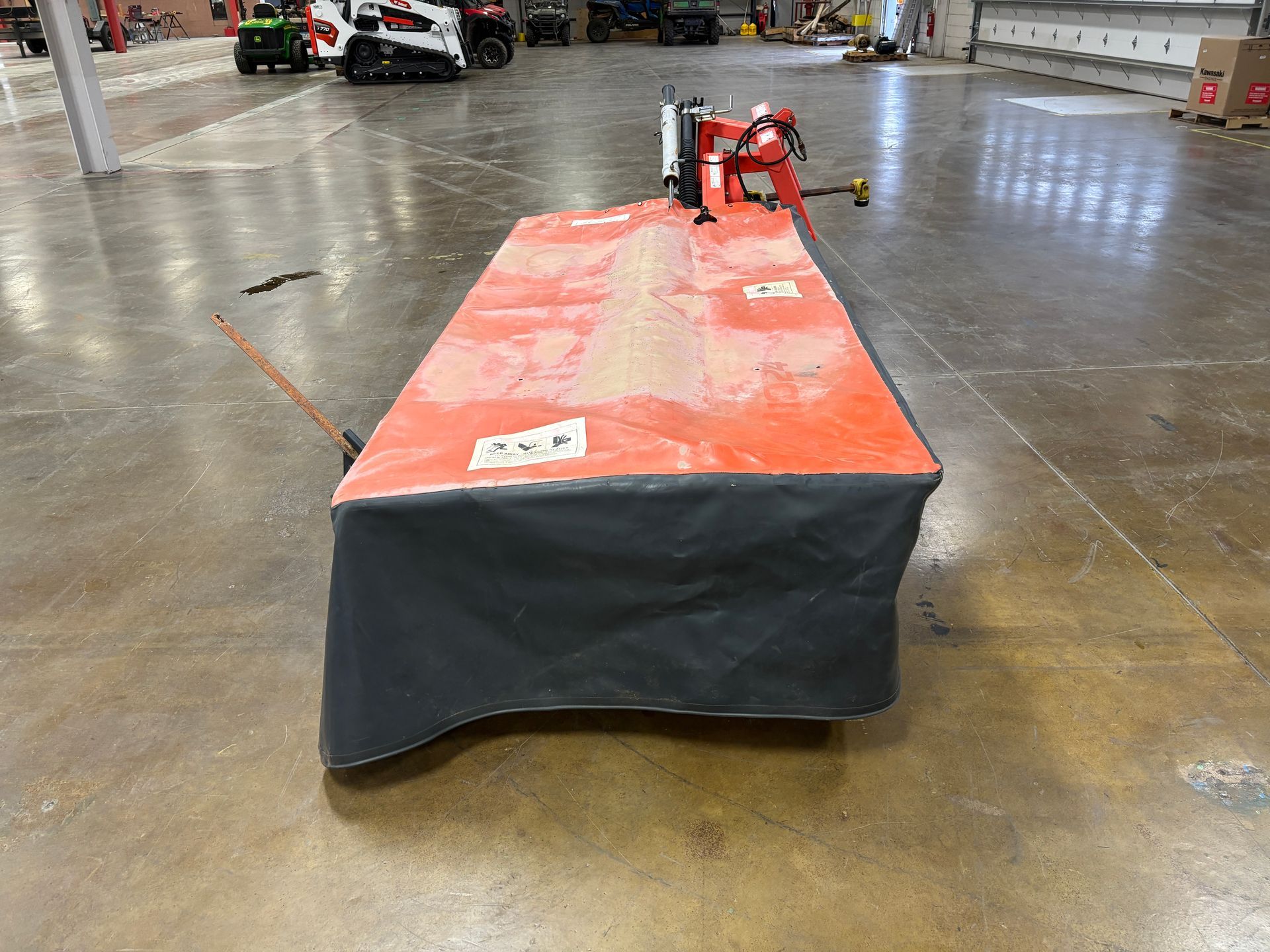 Kubota disc mower, in a orange and black cover, on a concrete floor, side view.