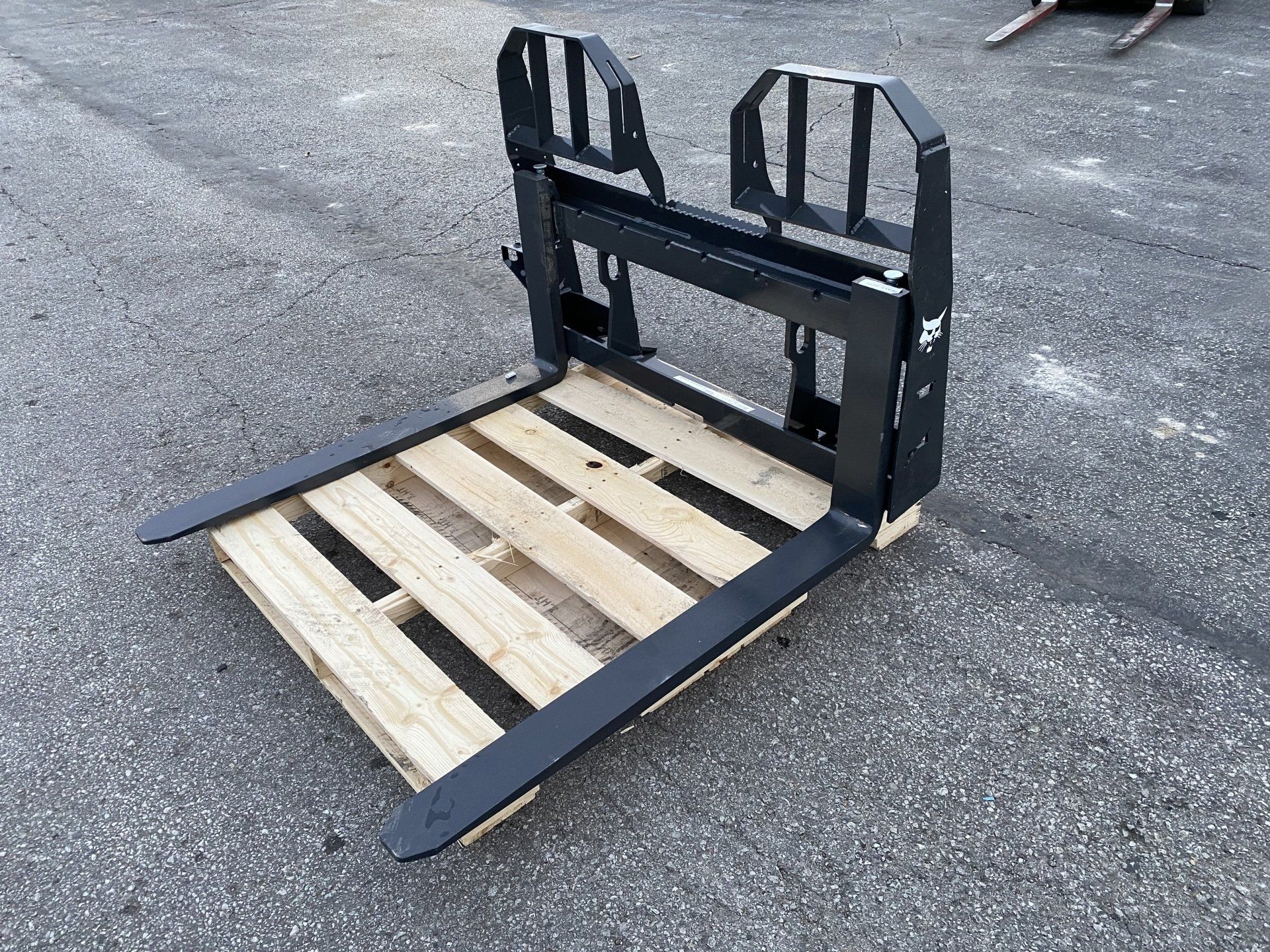 Black forklift attachment resting on a wooden pallet outdoors on gray asphalt.
