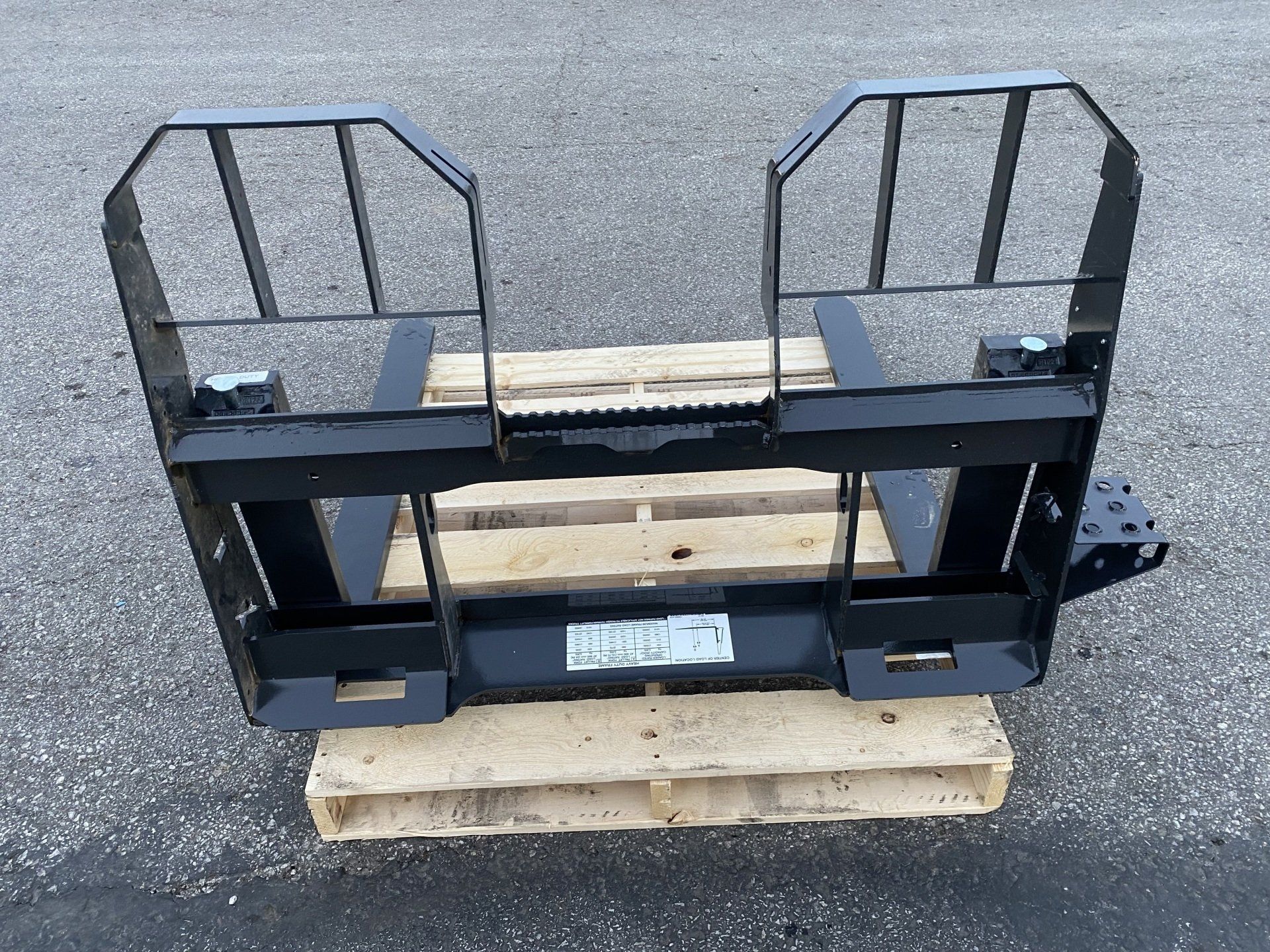 Black forklift attachment on a wooden pallet, with two fork guides and safety cages.
